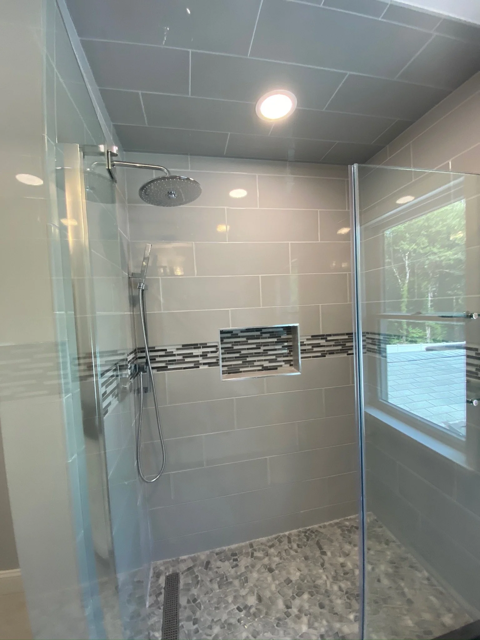 Modern shower with glass door, gray tiles, decorative tile strip, niche, rainfall showerhead, handheld shower, and window.