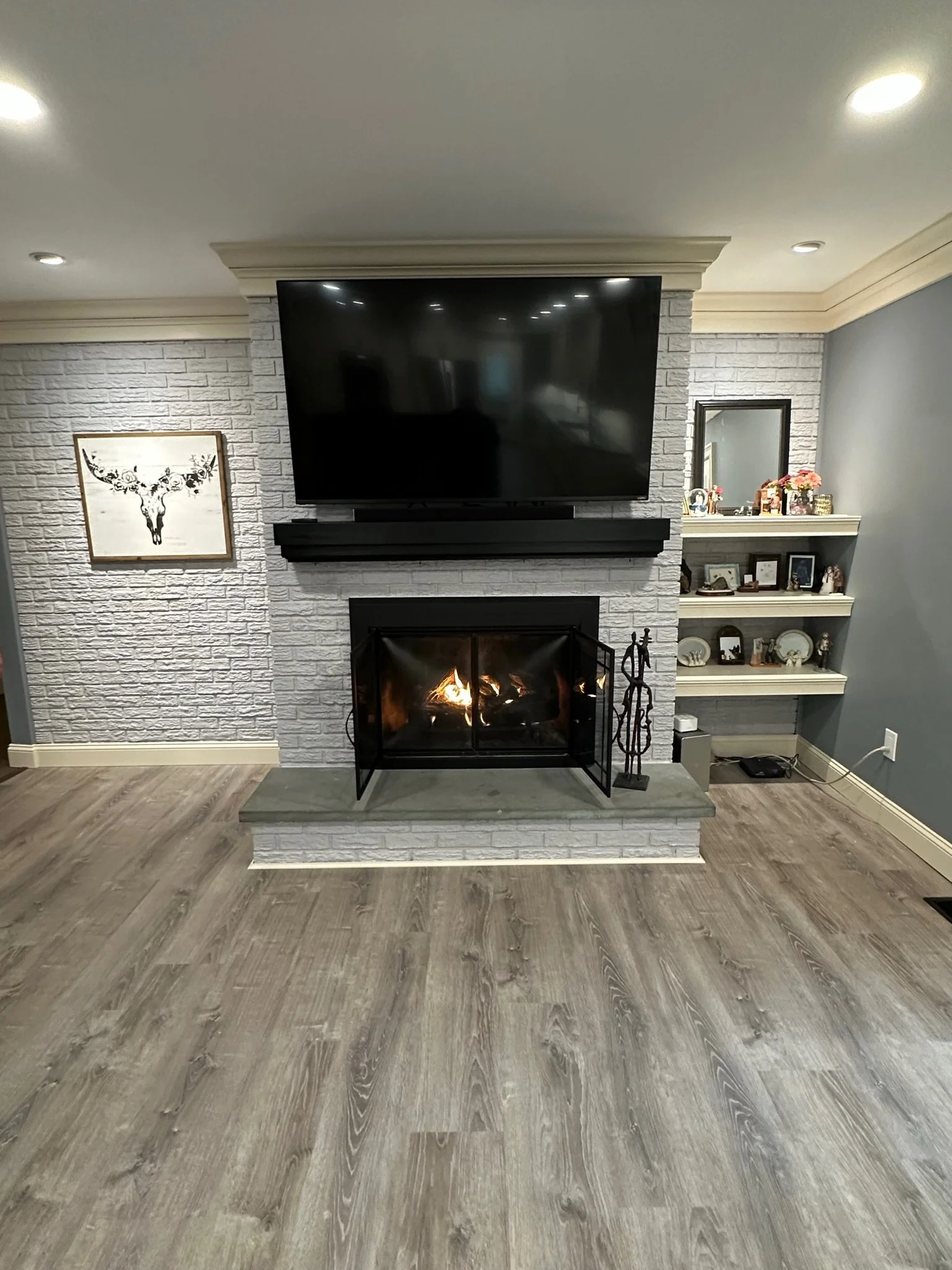 Living room with a stone fireplace, a television mounted above, and a wood floor. The fireplace has a fire burning and a decorative metal screen in front of it. To the right of the fireplace are wall shelves with decorations and photos. To the left i