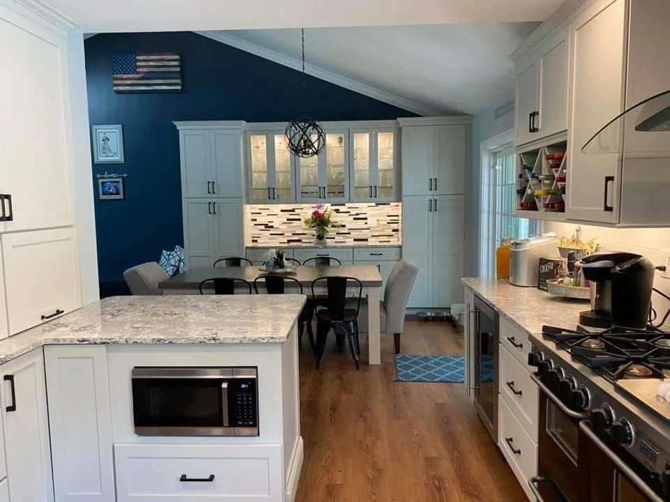 Open kitchen with white cabinets, granite countertops, gas stove, microwave, and coffee maker. Dining area with table, chairs, and a floral centerpiece. Decorated blue wall with American flag and framed artwork.