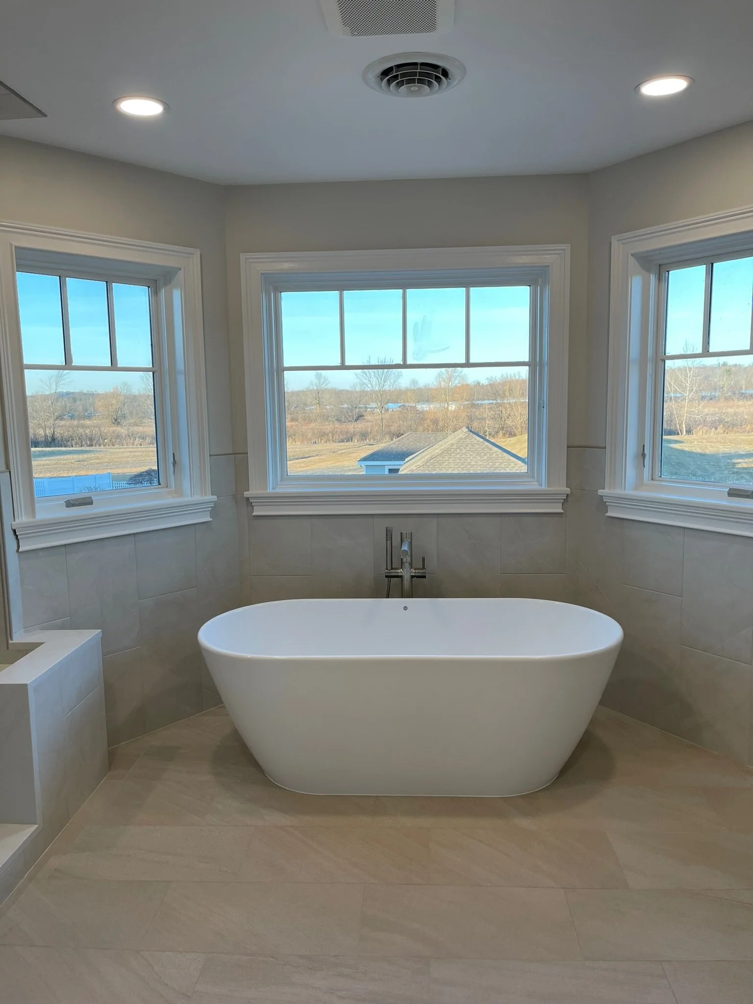 Modern bathroom with a white freestanding bathtub positioned in front of three large windows showing a rural landscape with fields and trees.