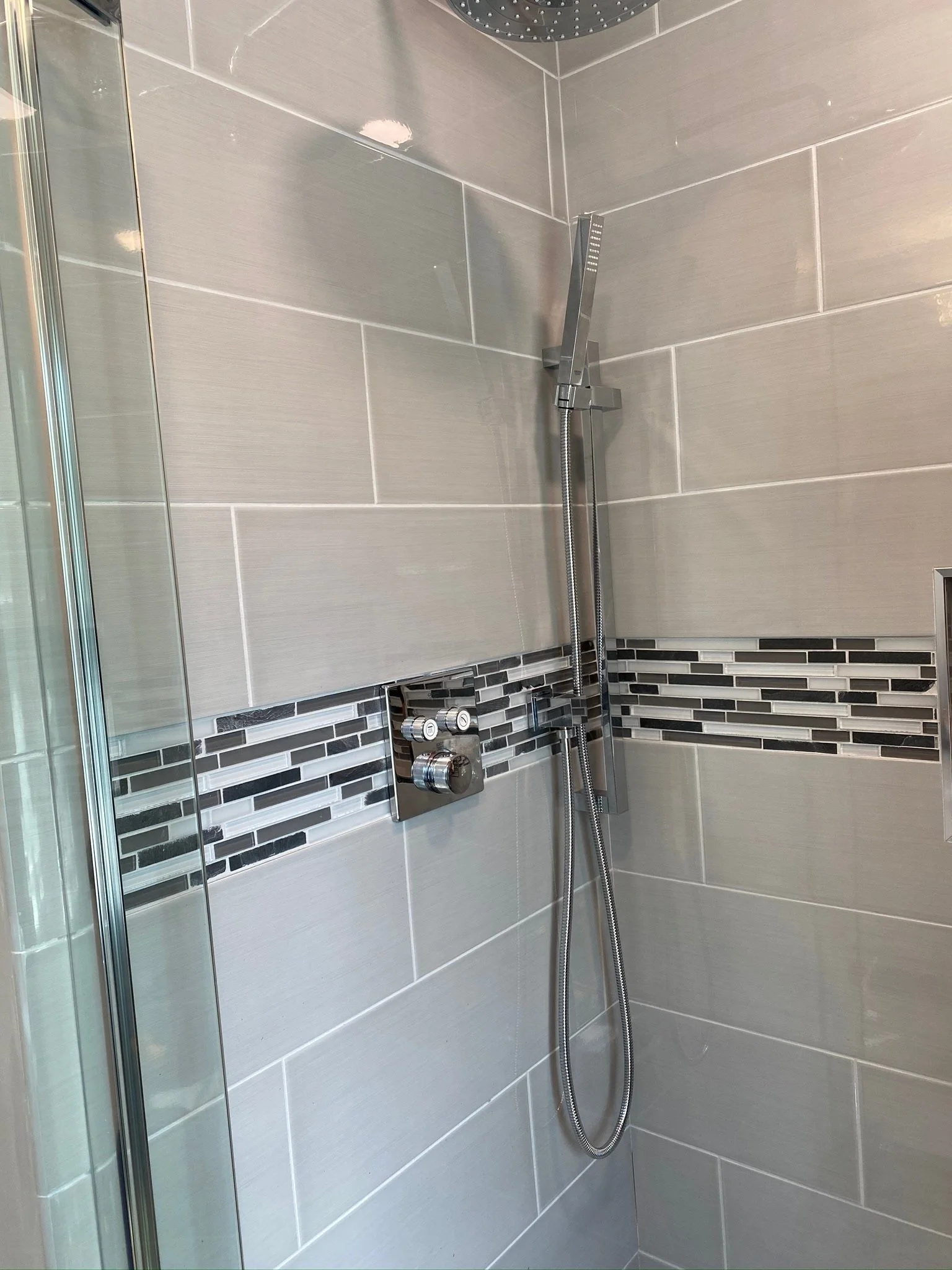 Shower with a large round rainfall head, a modern adjustable handheld shower, and a control panel with three knobs, set in a tiled bathroom wall with gray and black mosaic accents.