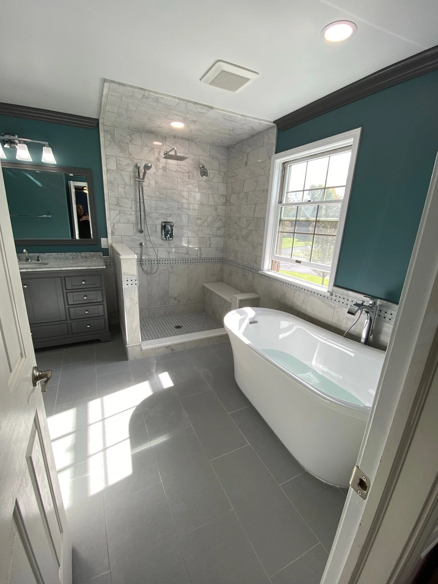 Bathroom with a freestanding bathtub, large window, gray vanity with mirror, and a walk-in shower with marble tiles and multiple showerheads.