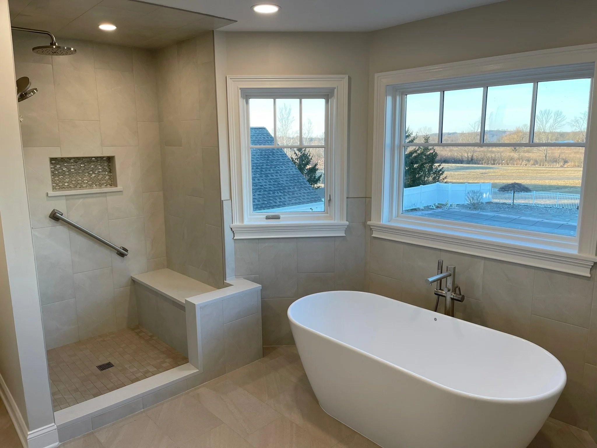 Modern bathroom featuring a walk-in shower with a rainfall showerhead, a built-in bench, and a white freestanding bathtub under large windows with a view of a backyard and open landscape.