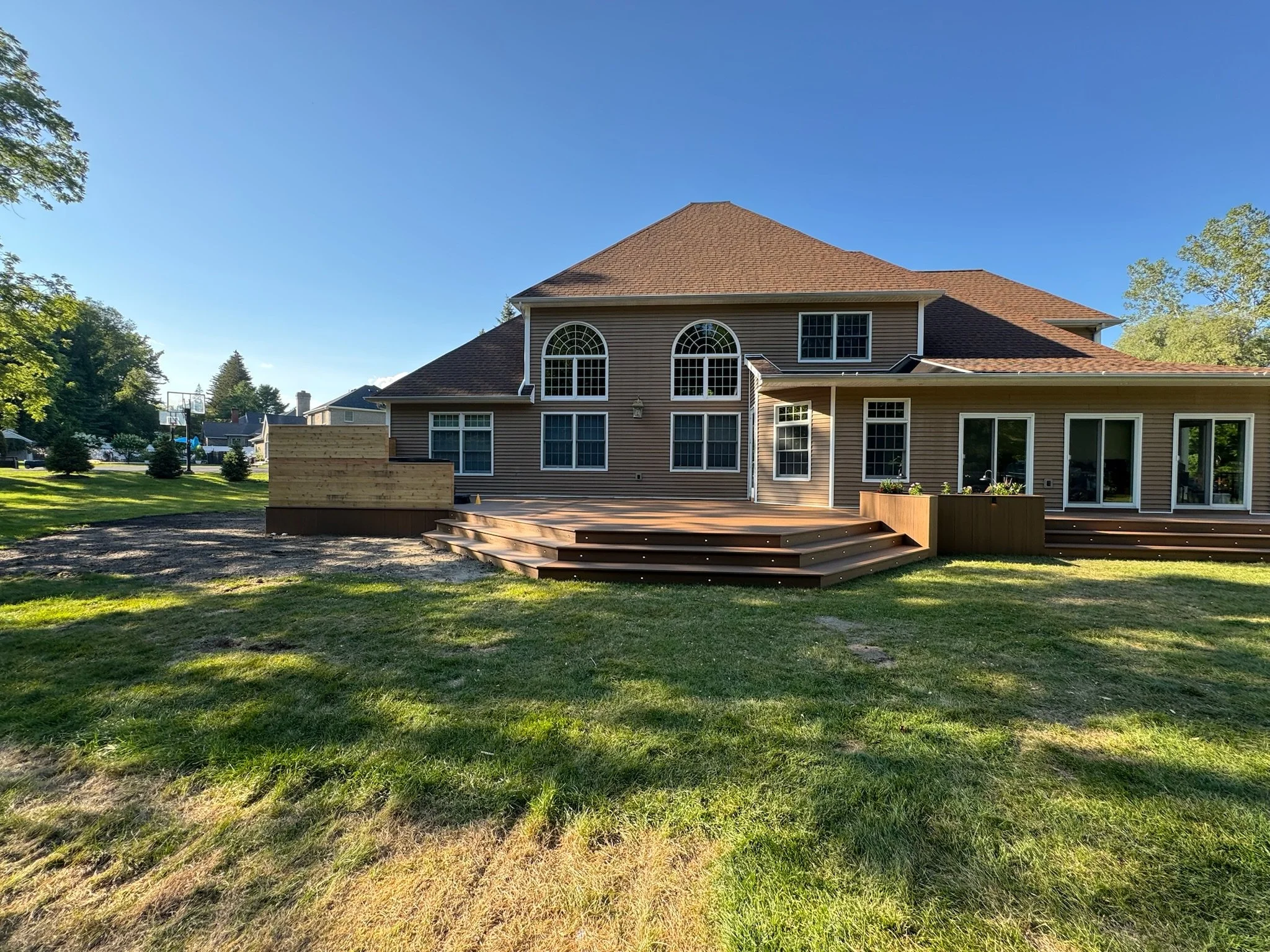 Backyard view of a two-story house with brown siding, a large wooden deck, and a grassy lawn under a clear blue sky.