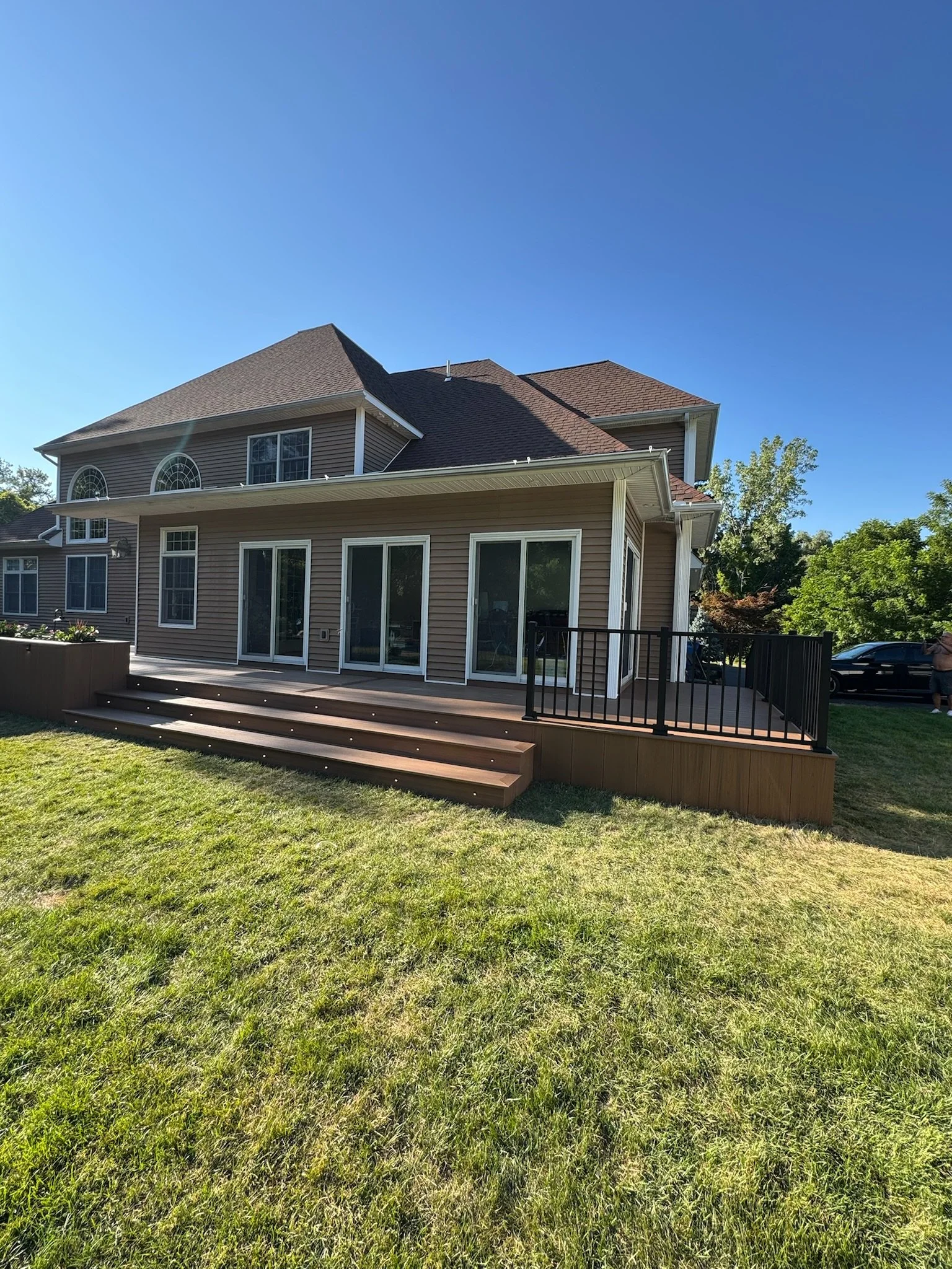 Backyard view of a two-story house with a wooden deck and sliding glass doors, surrounded by green grass and trees under a clear blue sky.