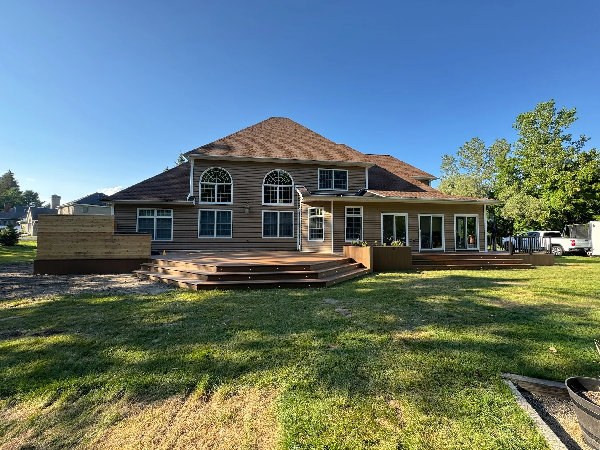 Back view of a large two-story house with a wooden deck and green lawn, blue sky, and trees in the background.