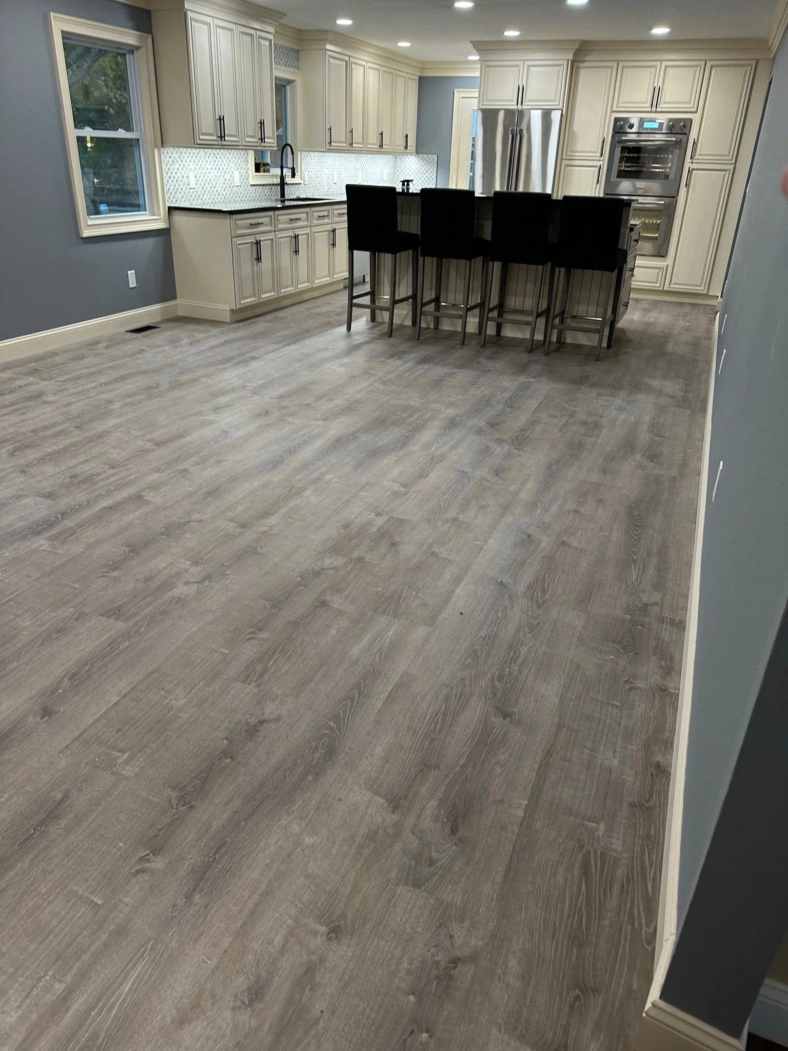 A spacious kitchen with white cabinetry, stainless steel appliances, a large island with black chairs, a gray backsplash, and hardwood flooring. Two windows are visible, providing natural light.