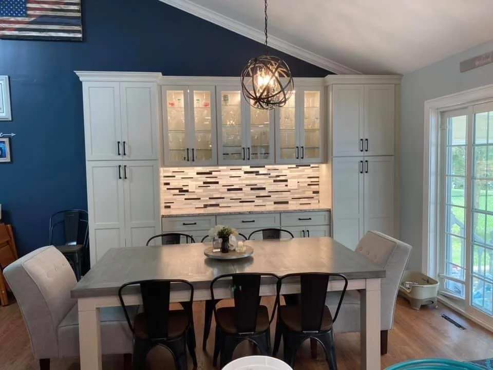 A dining area with a table surrounded by a mix of white and black chairs, a cabinet with glass doors and white cabinets, a modern light fixture, a window with a view of a green outdoor space, and a backsplash with a brick pattern.