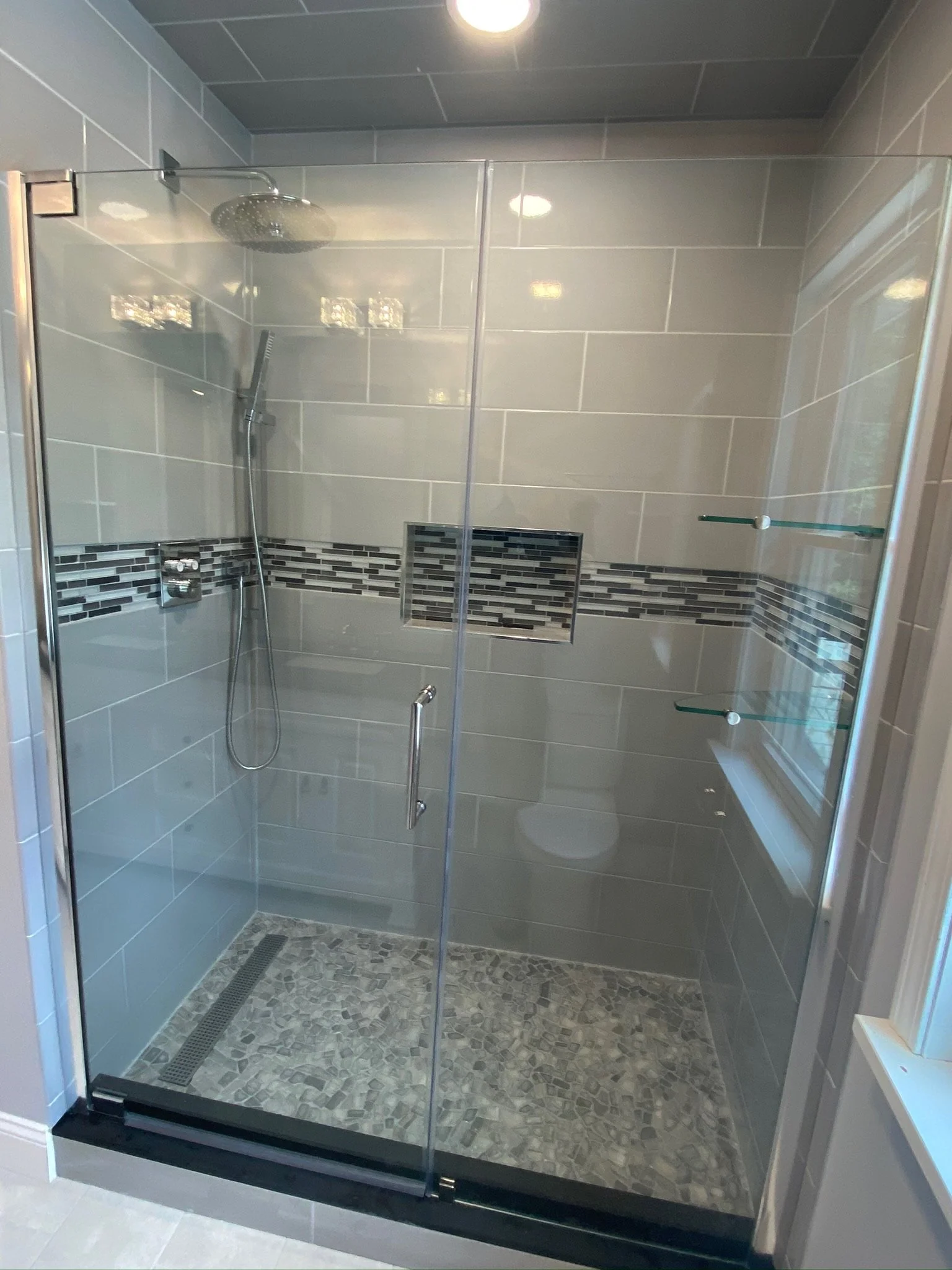 A modern glass-enclosed shower with beige tiles, a built-in niche with mosaic tile accents, a rainfall showerhead, a handheld showerhead, and glass shelves on the right side. There is also a window on the right wall.