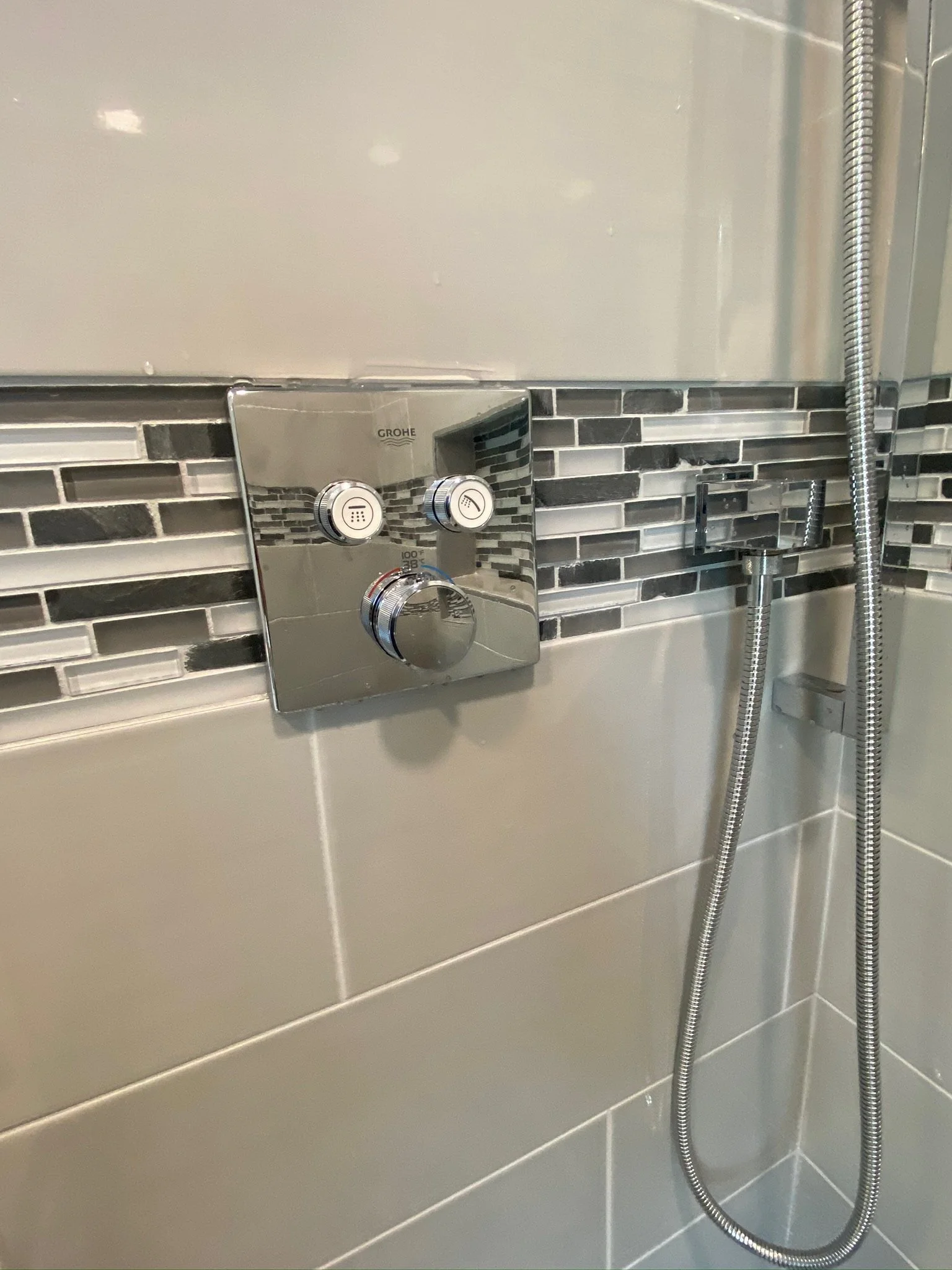 Close-up of a shower control panel with two knobs and one dial, mounted on a tiled bathroom wall with gray and black mosaic tiles.