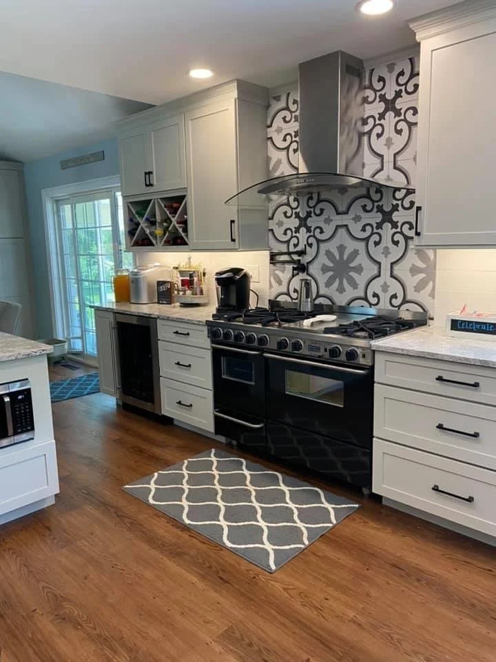 Modern kitchen with white cabinets, black appliances, patterned backsplash, wooden floor, gray rug, and a sliding glass door in the background.