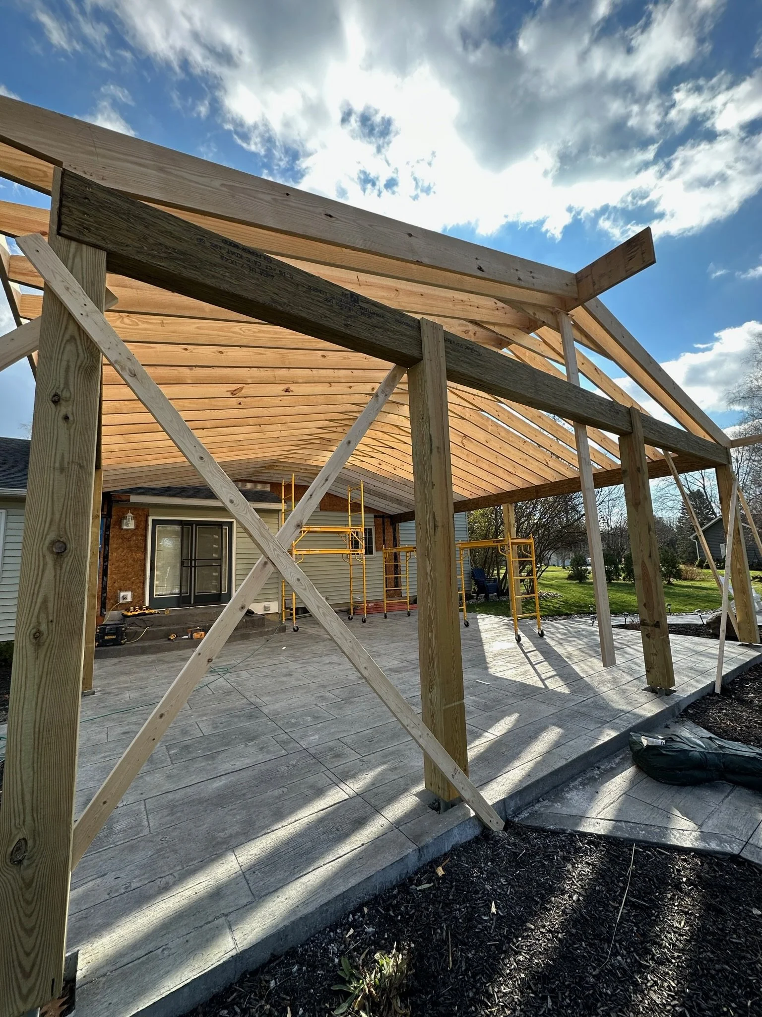 Construction site with a wooden framework being built over a patio, with blue sky and clouds overhead.