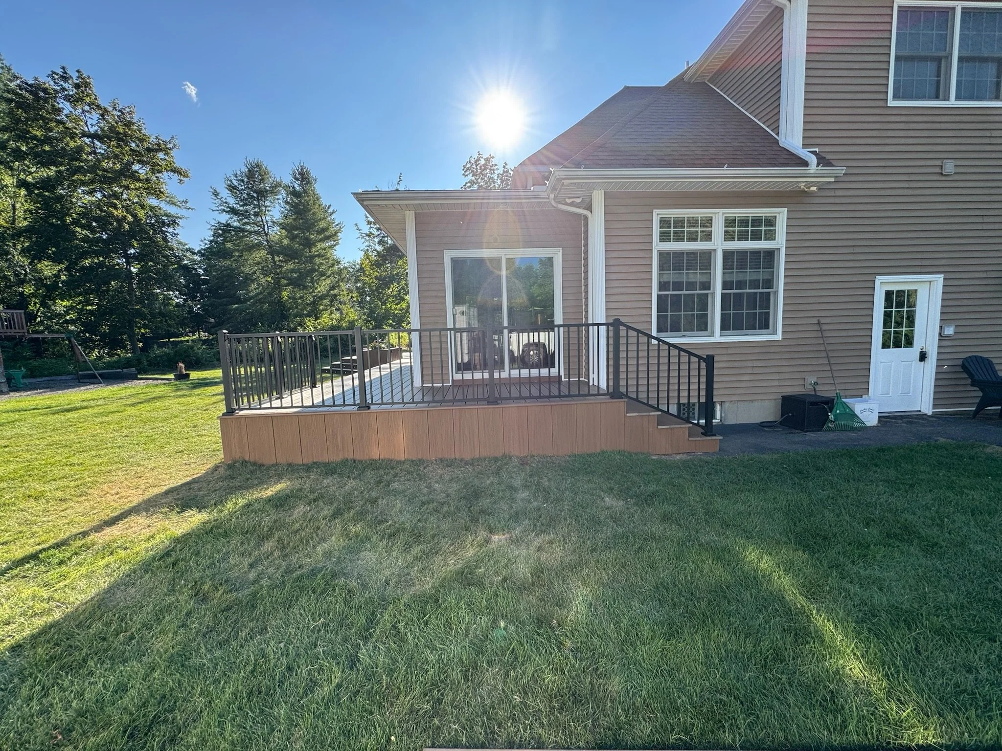 Backyard with a newly built wooden deck with black metal railings attached to a house. The house has tan siding, white trim, and multiple windows. The sun is shining brightly in a clear blue sky, casting shadows on the grass and deck.