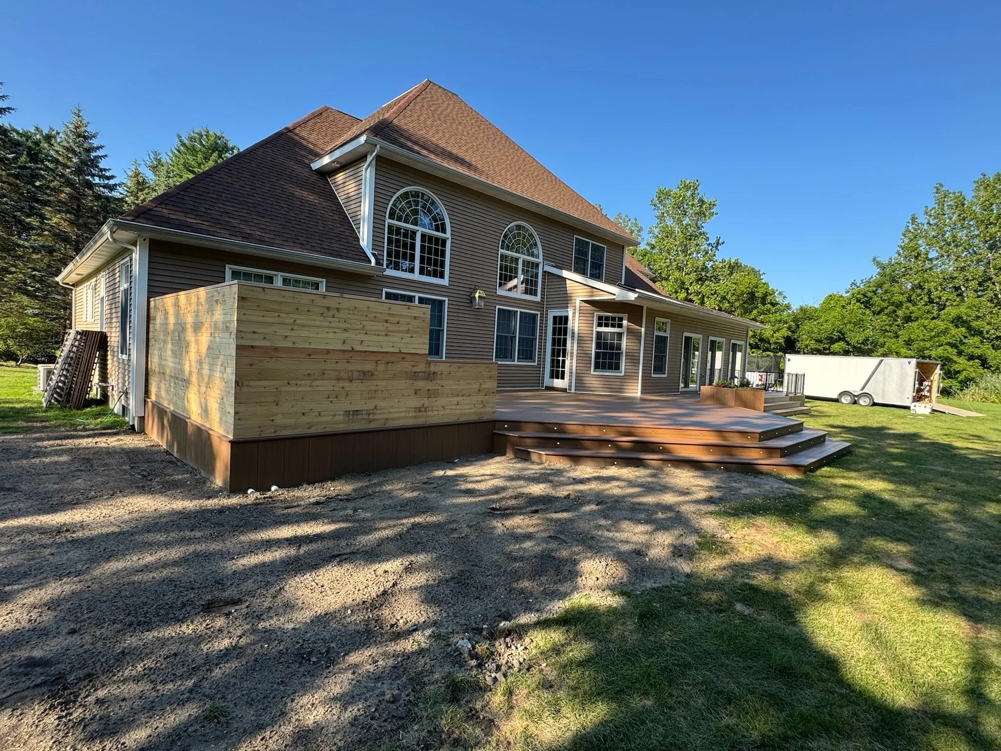 A two-story house with a brown roof and beige siding, featuring large arched windows in the upper story. There is a newly built wooden deck extending from the back of the house with a set of stairs leading down to the lawn. The yard appears to be und