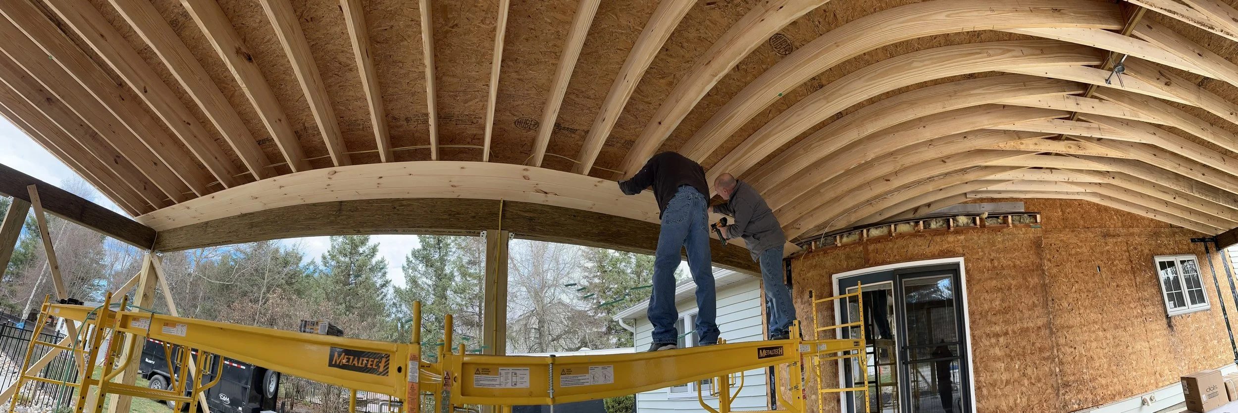 Two construction workers installing a curved wooden support beam under a roof extension of a house, with framing and plywood visible, using a scissor lift.