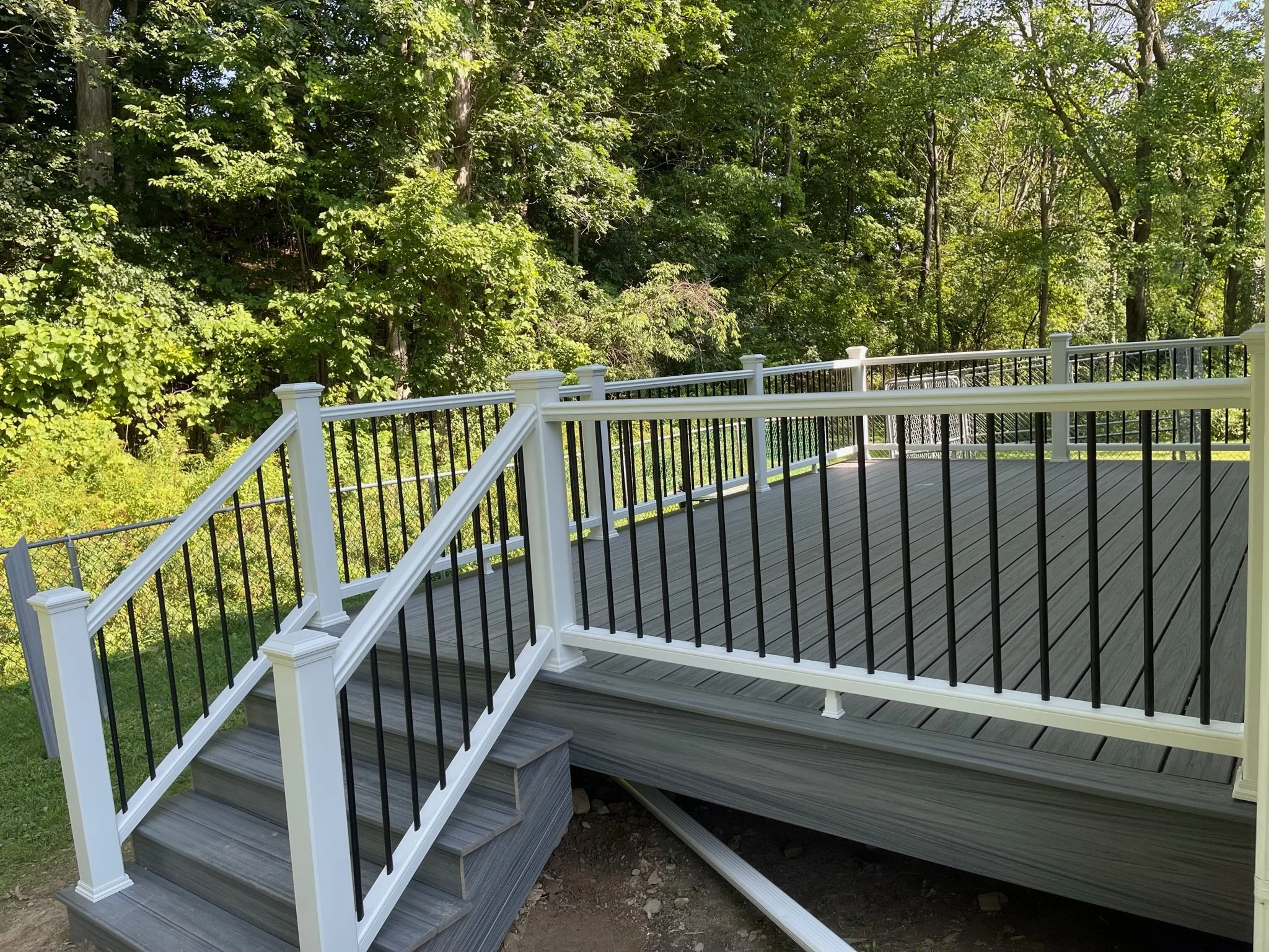 A wooden deck with black metal railing and stairs leading down to the ground, surrounded by lush green trees.