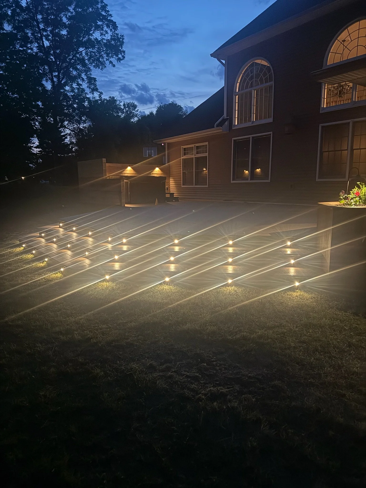 A backyard with a patio and house illuminated by outdoor lighting at dusk, featuring a large tree and a volleyball net in the background.