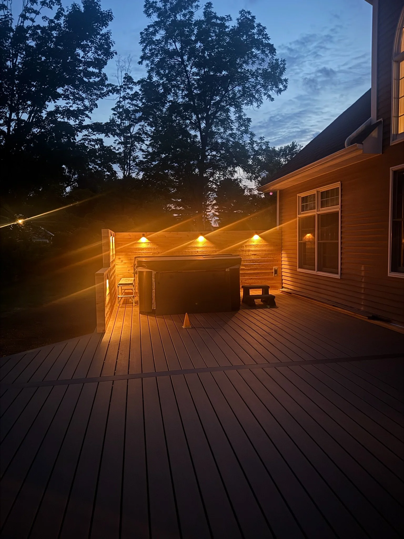 Outdoor wooden deck with a hot tub, small black step stool, and a privacy fence illuminated by warm outdoor lights during dusk, with trees and a cloudy sky in the background.