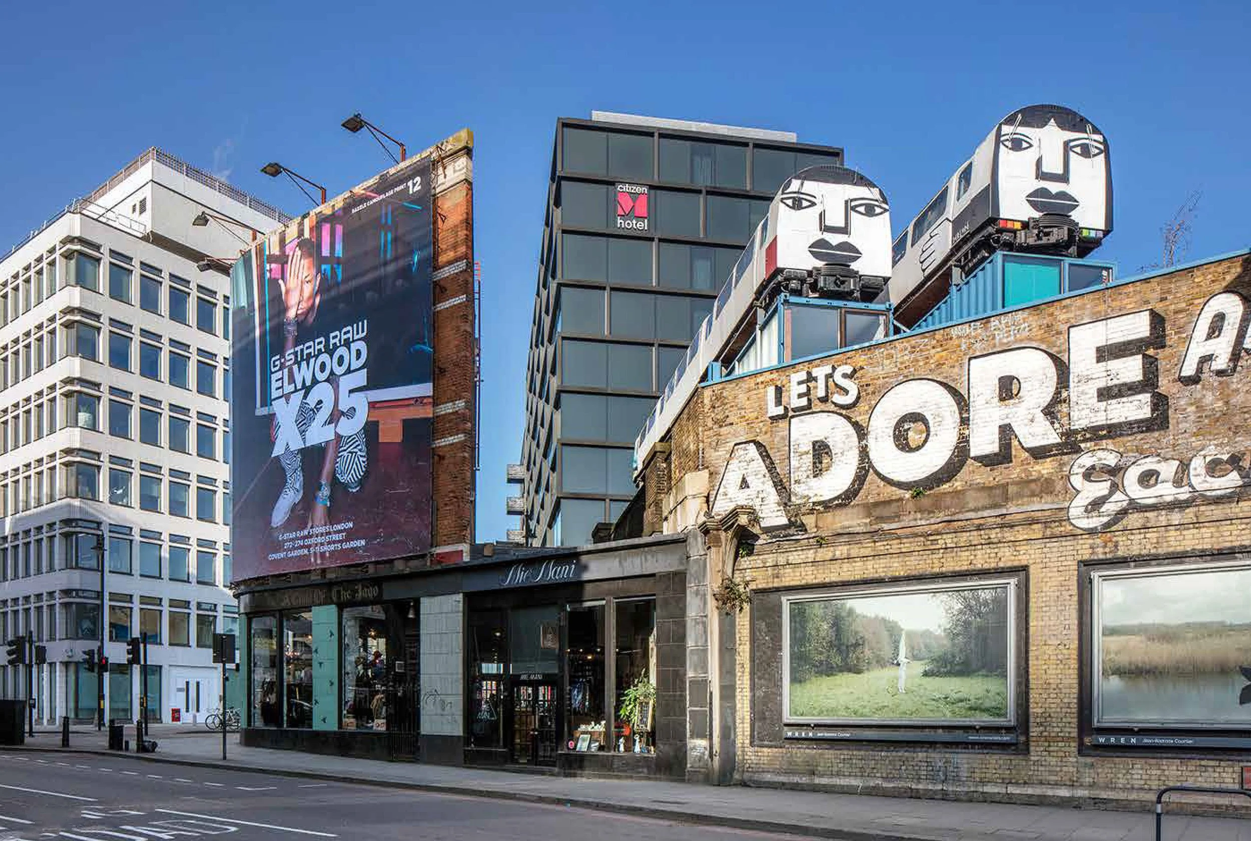 Urban street scene with tall buildings, billboard with G-Star RAW advertisement, two large female face sculptures on the roof of a building, and a sign reading "LET'S ADORE."