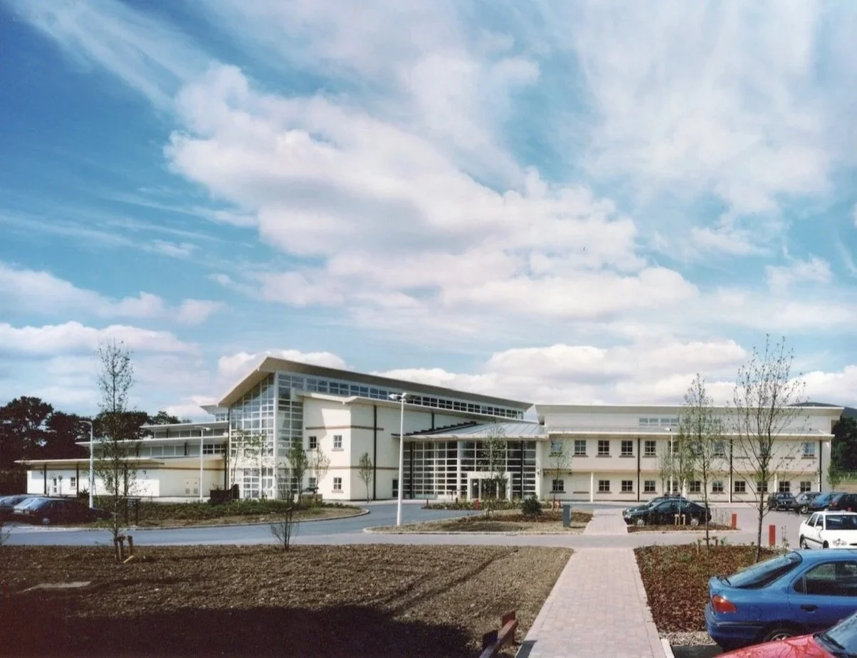 Modern building with glass windows and a parking lot, surrounded by trees under a partly cloudy sky.