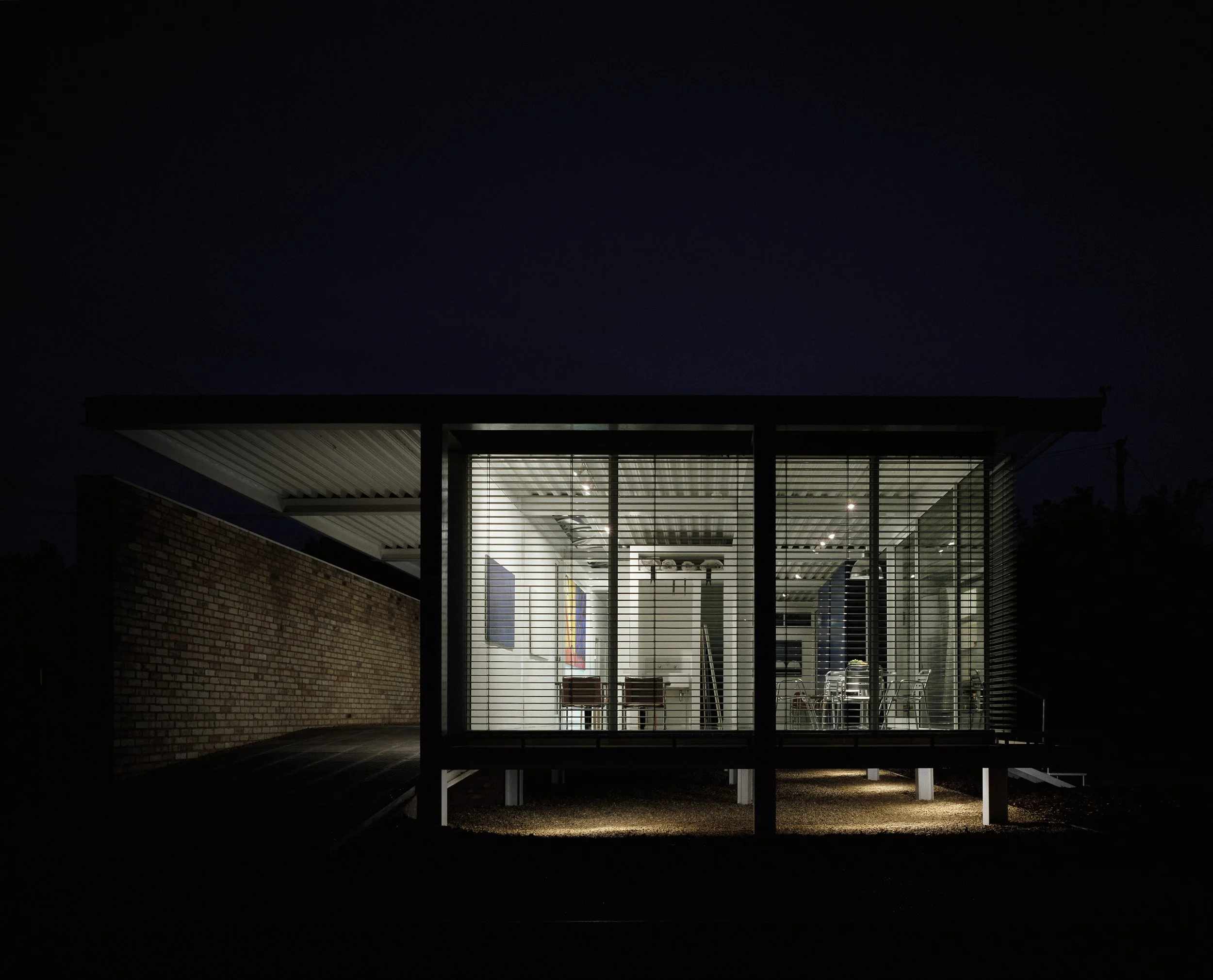 Modern glass-walled house at night with interior lighting showing a dining area and kitchen, surrounded by darkness.