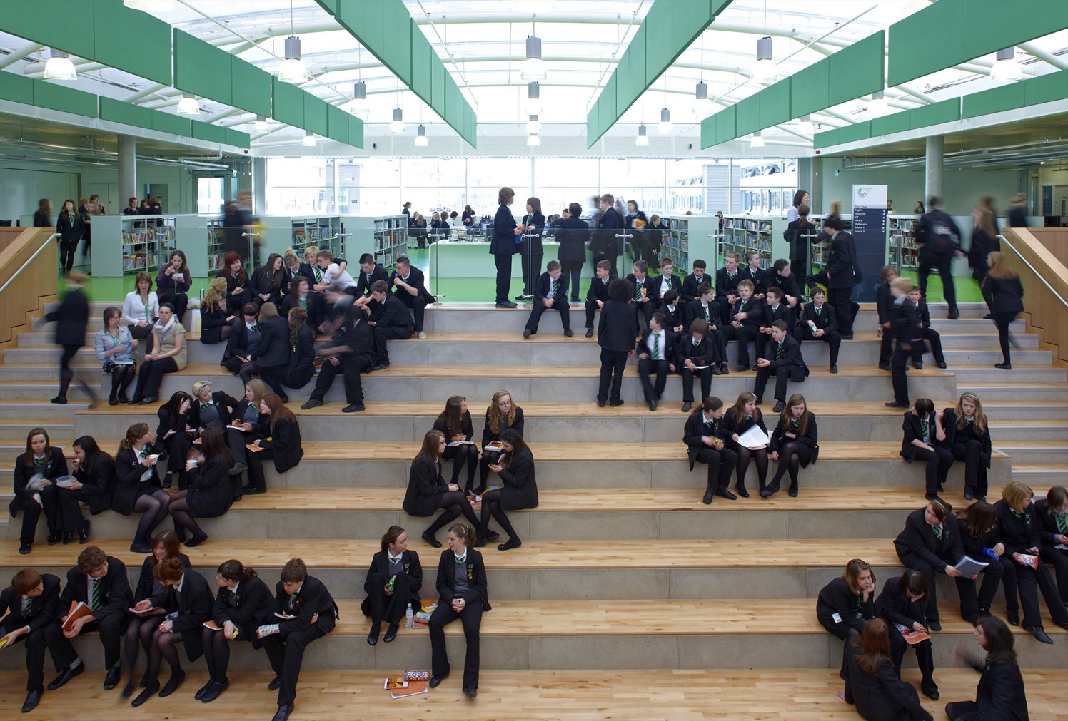 Ellis-Miller and Partners — Students dressed in school uniforms sitting on steps and standing in a modern library or educational space with bookshelves and large windows.