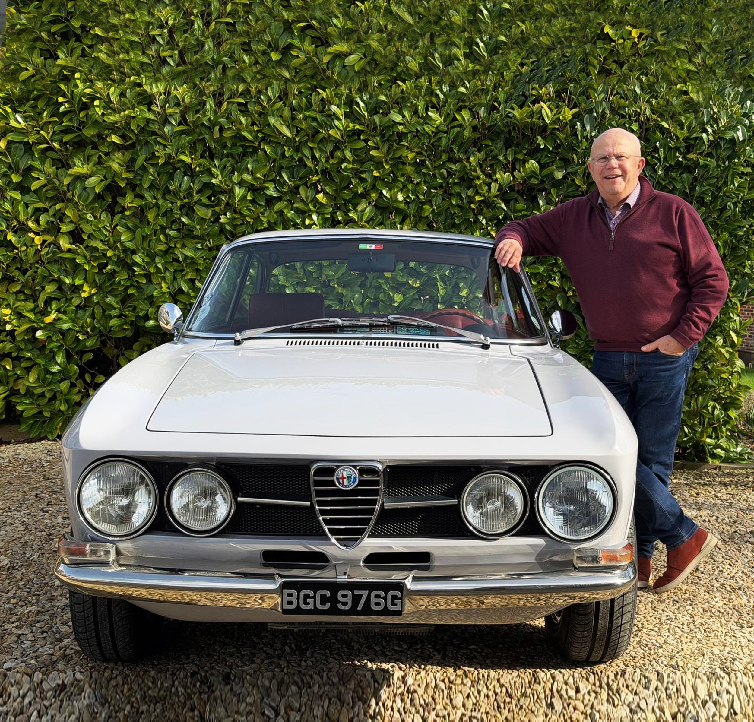 A man leaning on the door of a white vintage Alfa Romeo car parked on a gravel driveway with green bushes in the background.