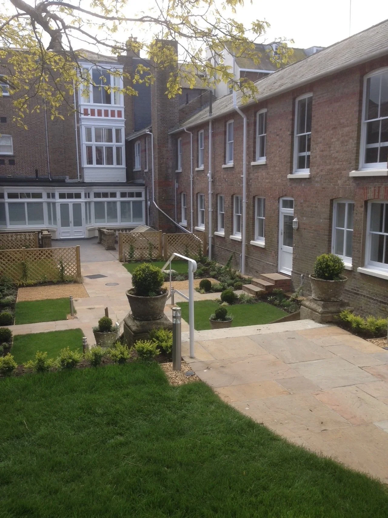 A courtyard with green grass, small plants, stone planters, and paved walkways surrounded by brick buildings with white-framed windows.