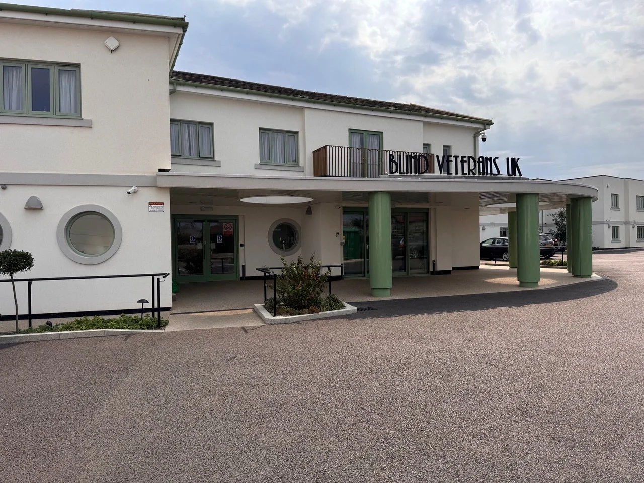 Front entrance of a building with a sign that reads 'Blind Veterans UK' on top, featuring green pillars and circular windows, parking area with cars in the background.