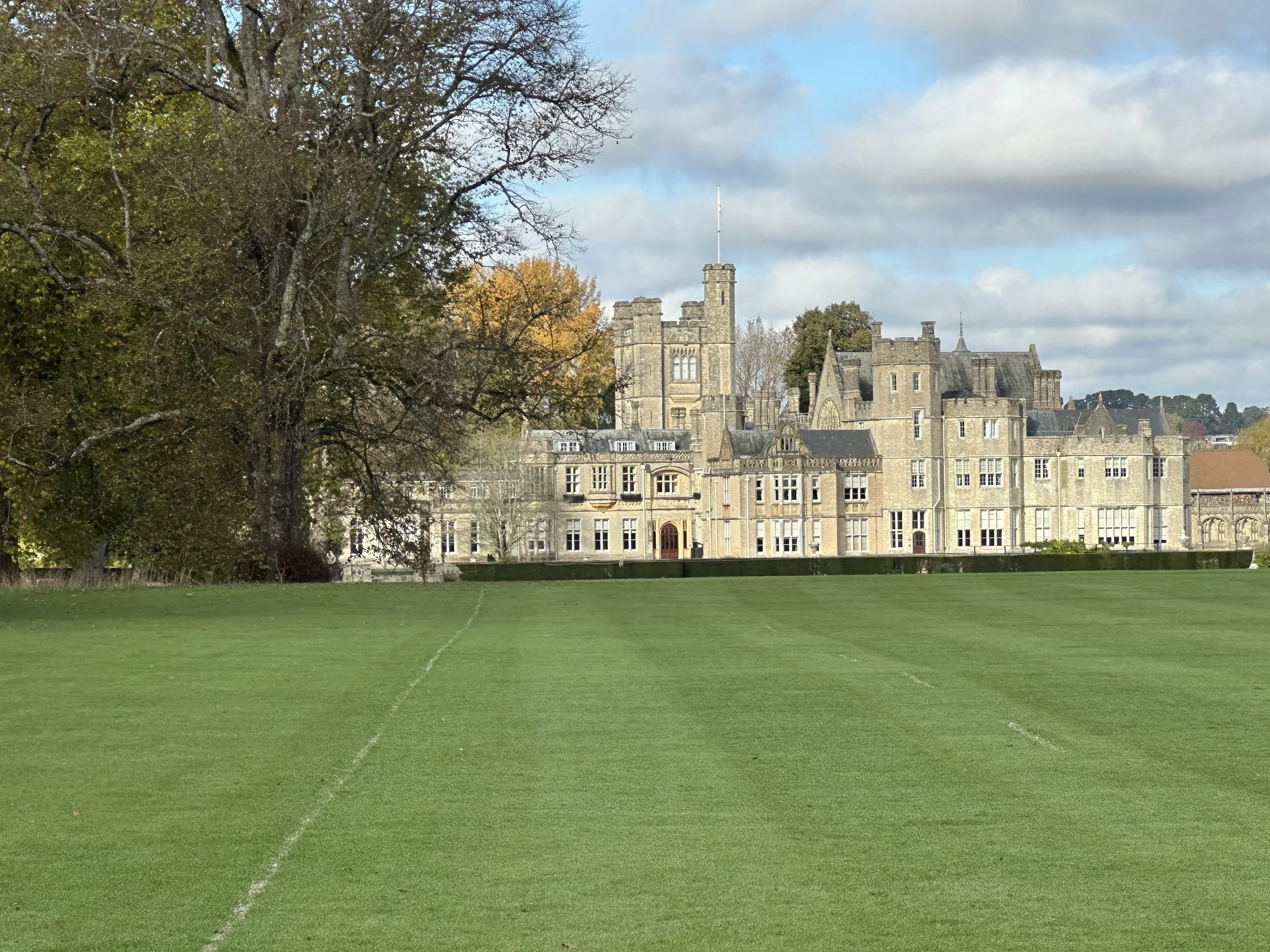 A large historic castle or mansion surrounded by a lawn and trees with autumn foliage, under a partly cloudy sky.