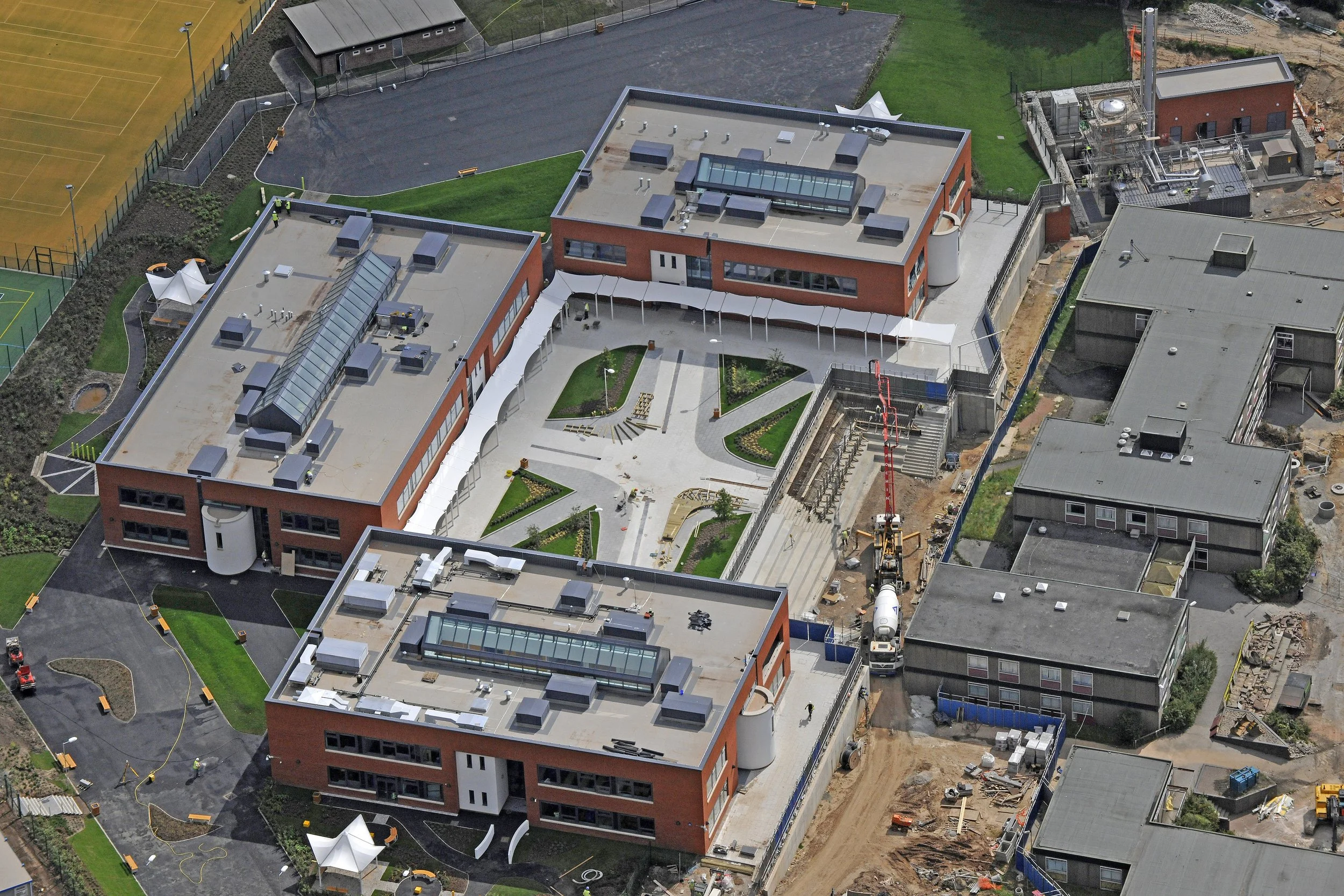 An aerial view of a modern school campus under construction, featuring multiple brick buildings, a courtyard with landscaped gardens and benches, sports fields, and construction equipment.