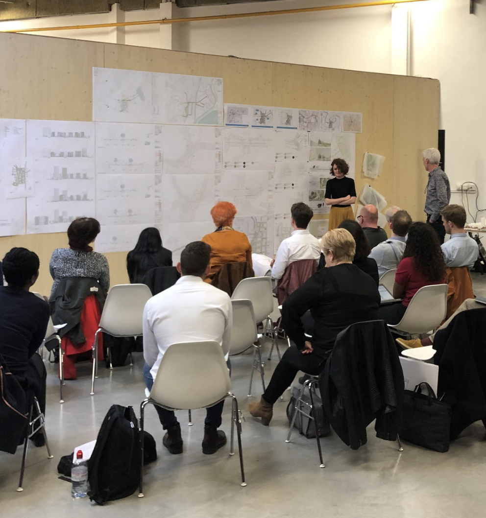 Group of people attending a presentation or seminar in a large room, sitting on white chairs, listening to a woman speaking in front of large printed posters on a beige wall.