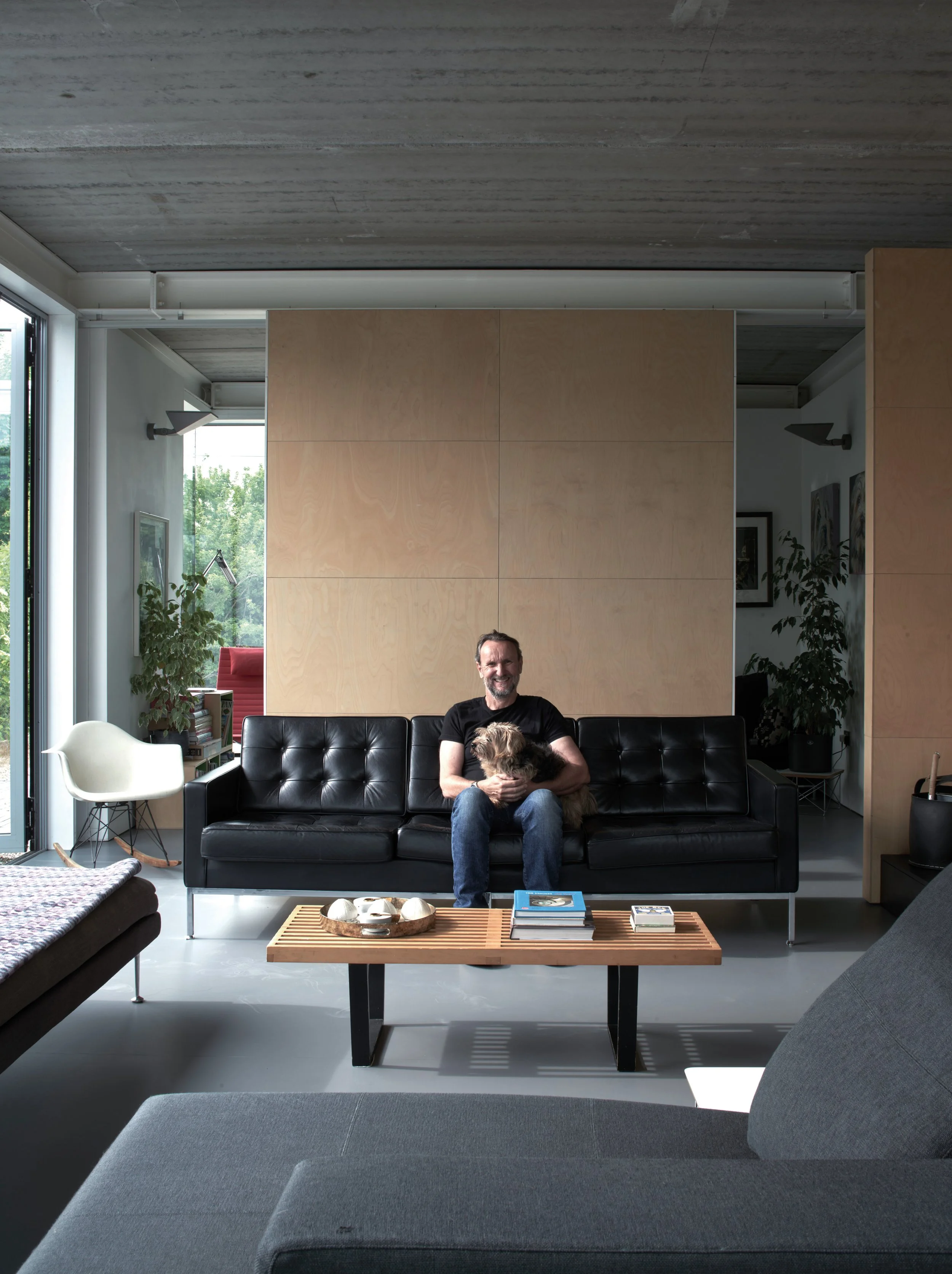 A man sitting on a black leather sofa holding a small dog, in a modern living room with a coffee table and various plants.
