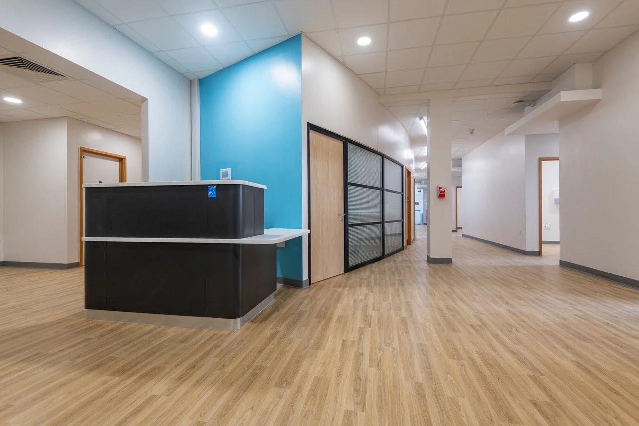 Interior of an office building with a reception desk, wooden flooring, white walls, and glass partitions.