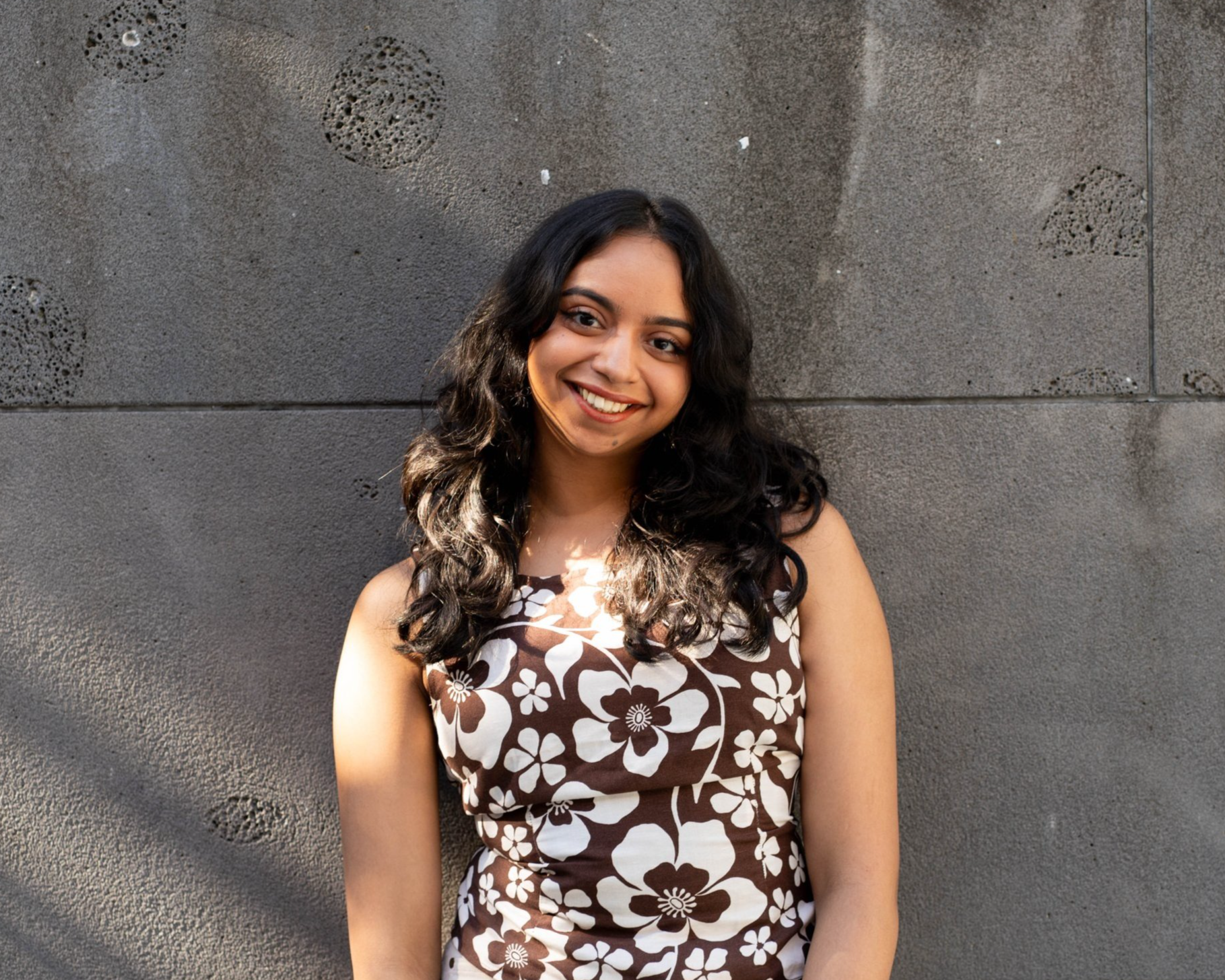 A young woman with long dark curly hair smiling, standing against a textured gray wall, wearing a sleeveless brown and white floral dress.