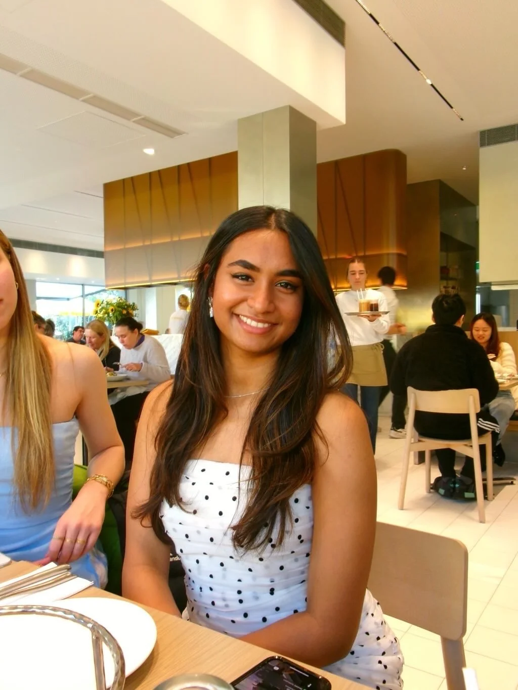A smiling young woman with long dark hair wearing a white dress with black polka dots, sitting at a restaurant table during the daytime.