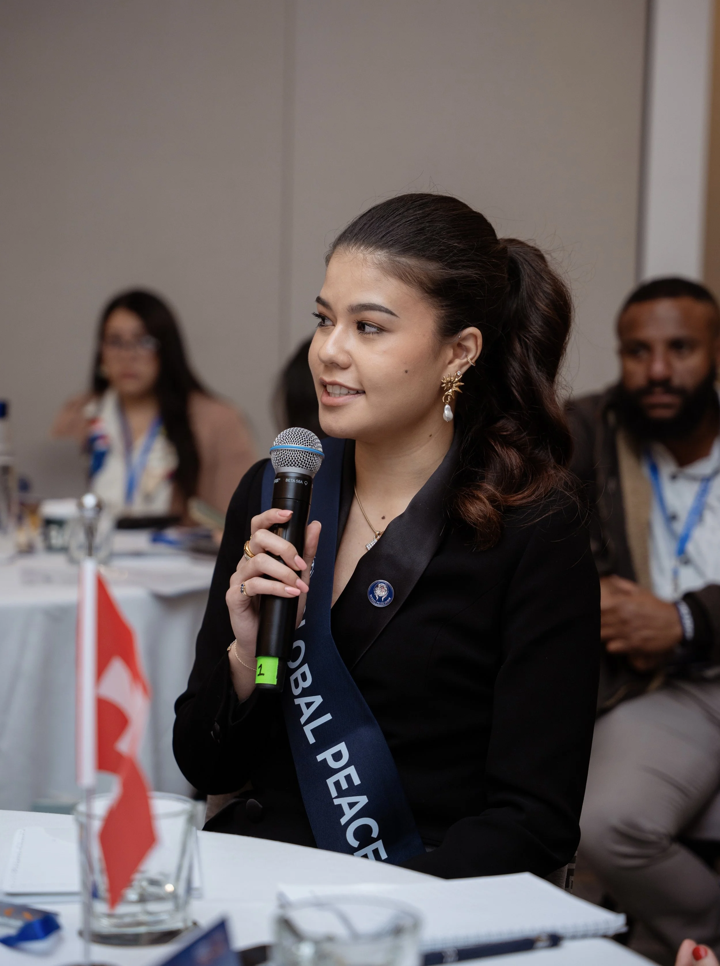 Young woman wearing a black blazer, pearl earring, holding a microphone, and wearing a sash that says "Global Peace" at a conference.
