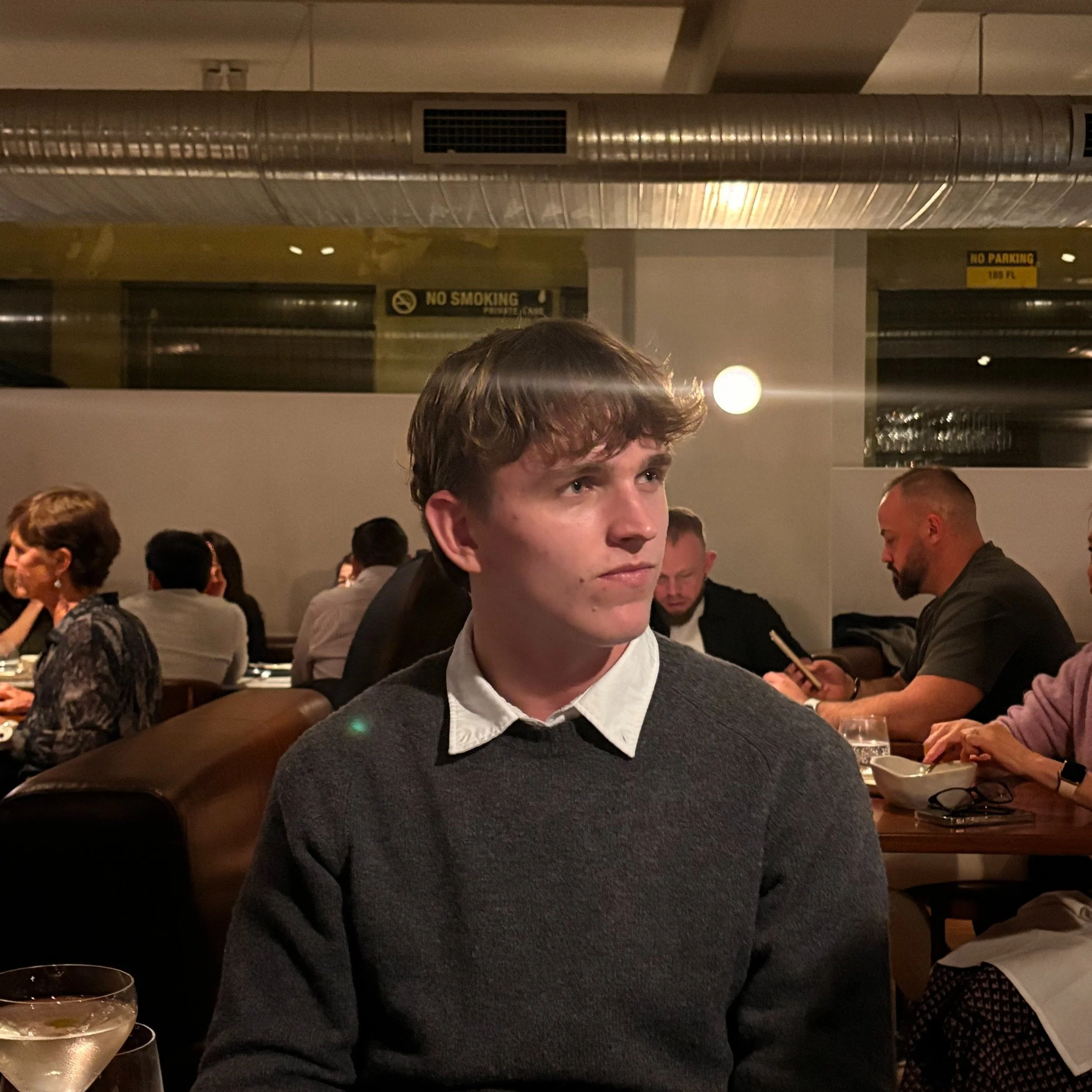 A young man with light brown hair wearing a white collared shirt under a dark sweater, sitting in a restaurant surrounded by other diners.