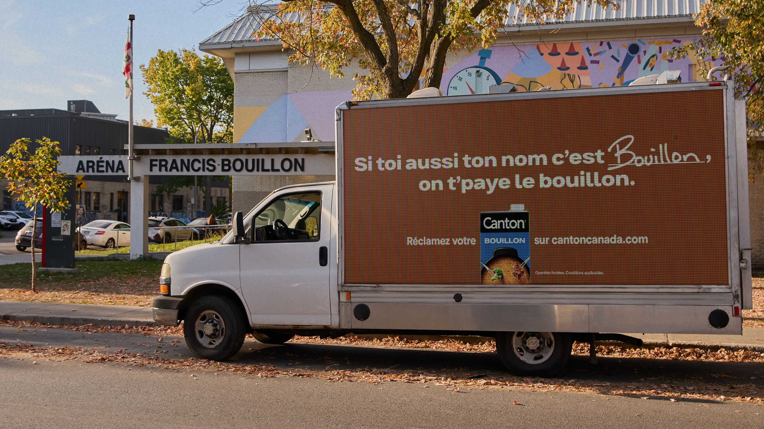 Un camion publicitaire garé devant un bâtiment nommé "Aréna Francis Bouillon" avec un arbre au premier plan, à l'automne.