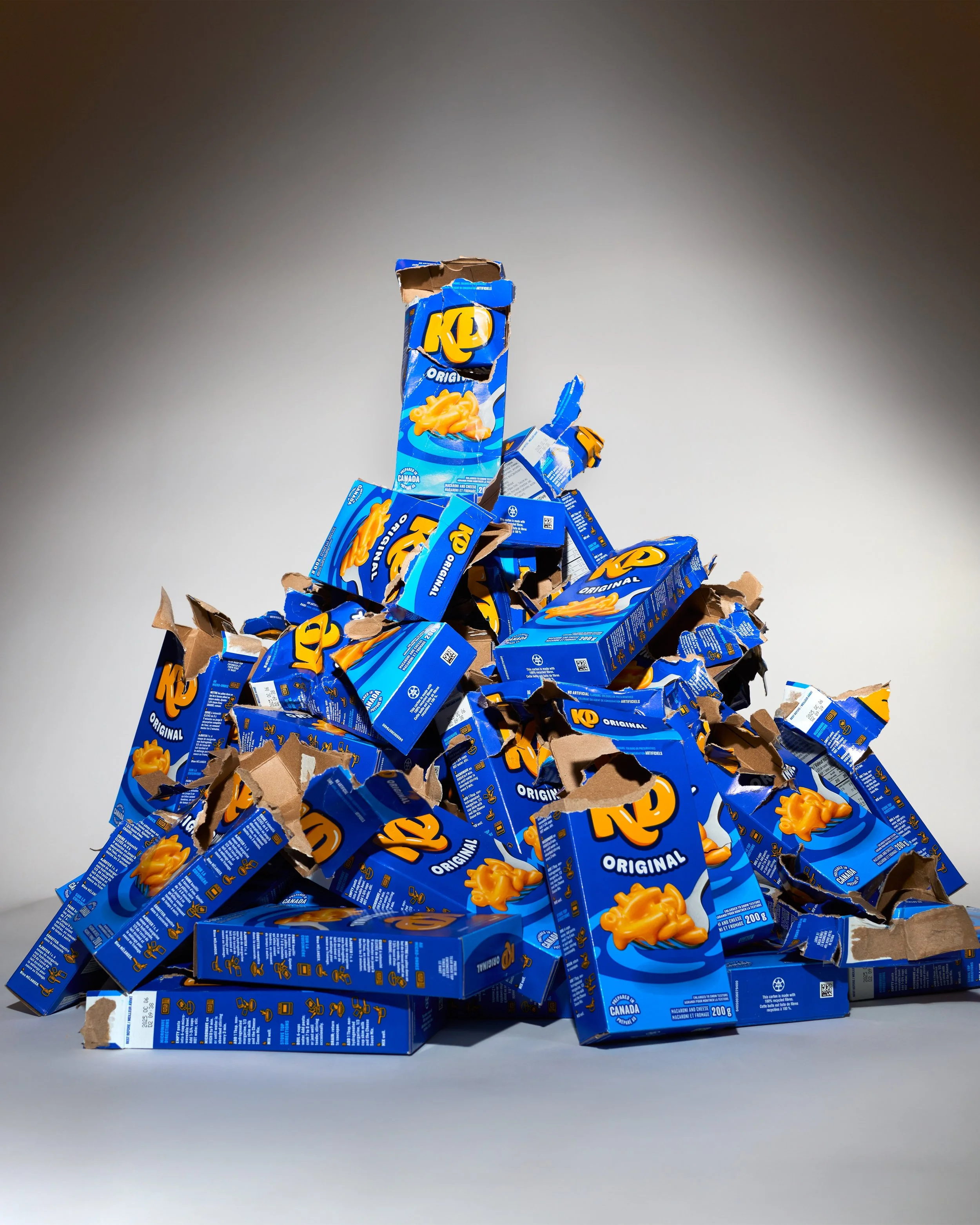 Monticule de boîtes de fromage cheddar Kraft en carton bleu, some ouvertes, quelques morceaux éparpillés, sur fond gris