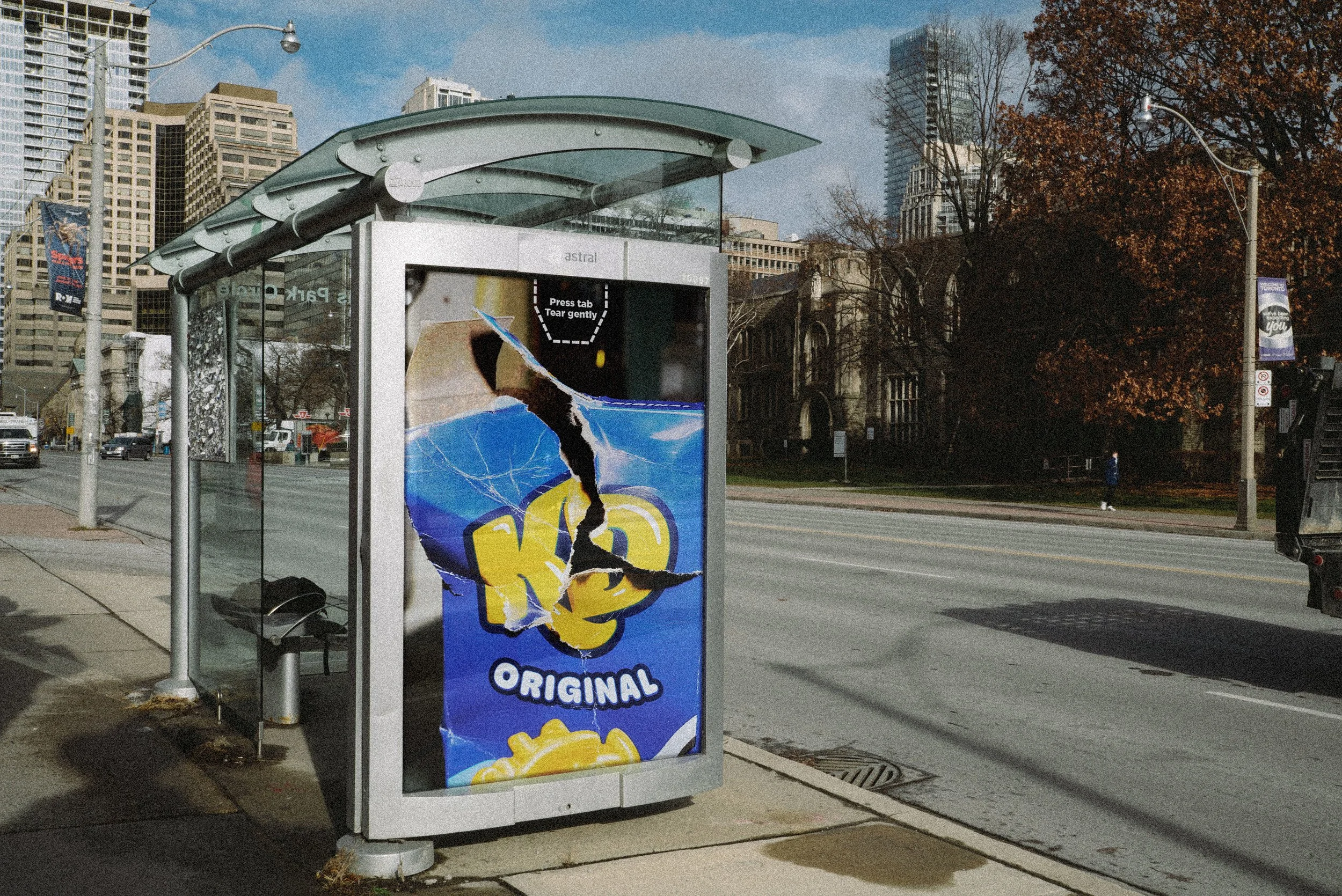 Un abri d'arrêt de bus avec une affiche déchirée de chips et un paysage urbain avec des immeubles, des arbres sans feuilles, une rue et un passage piéton.