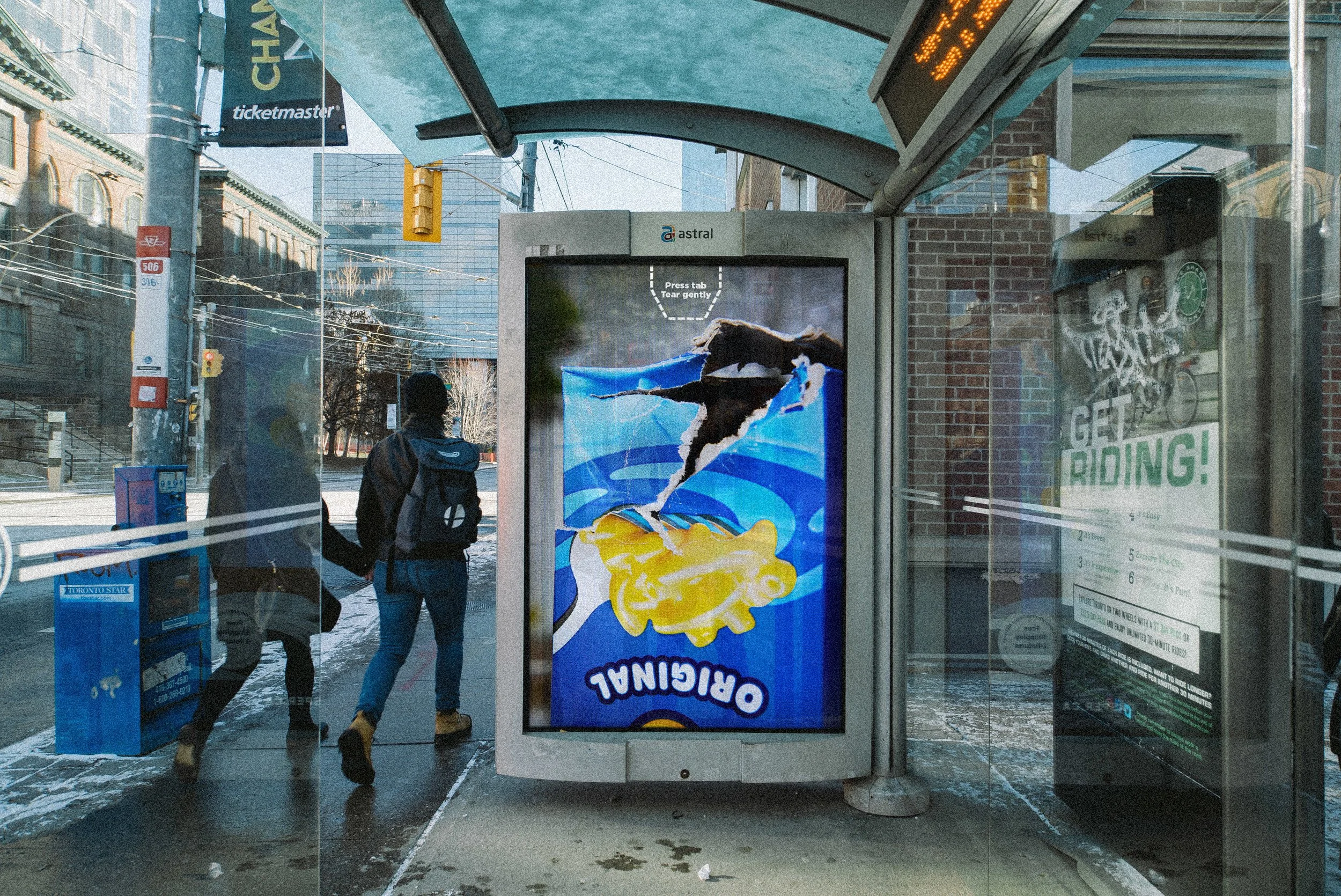 Une publicité sur un écran d'affichage urbain montre un bonhomme qui glisse sur une surface bleue, avec une tête en forme de croustilles. Deux personnes marchent devant l'affiche, dans un arrêt de bus à l'extérieur en hiver, avec de la neige au sol.