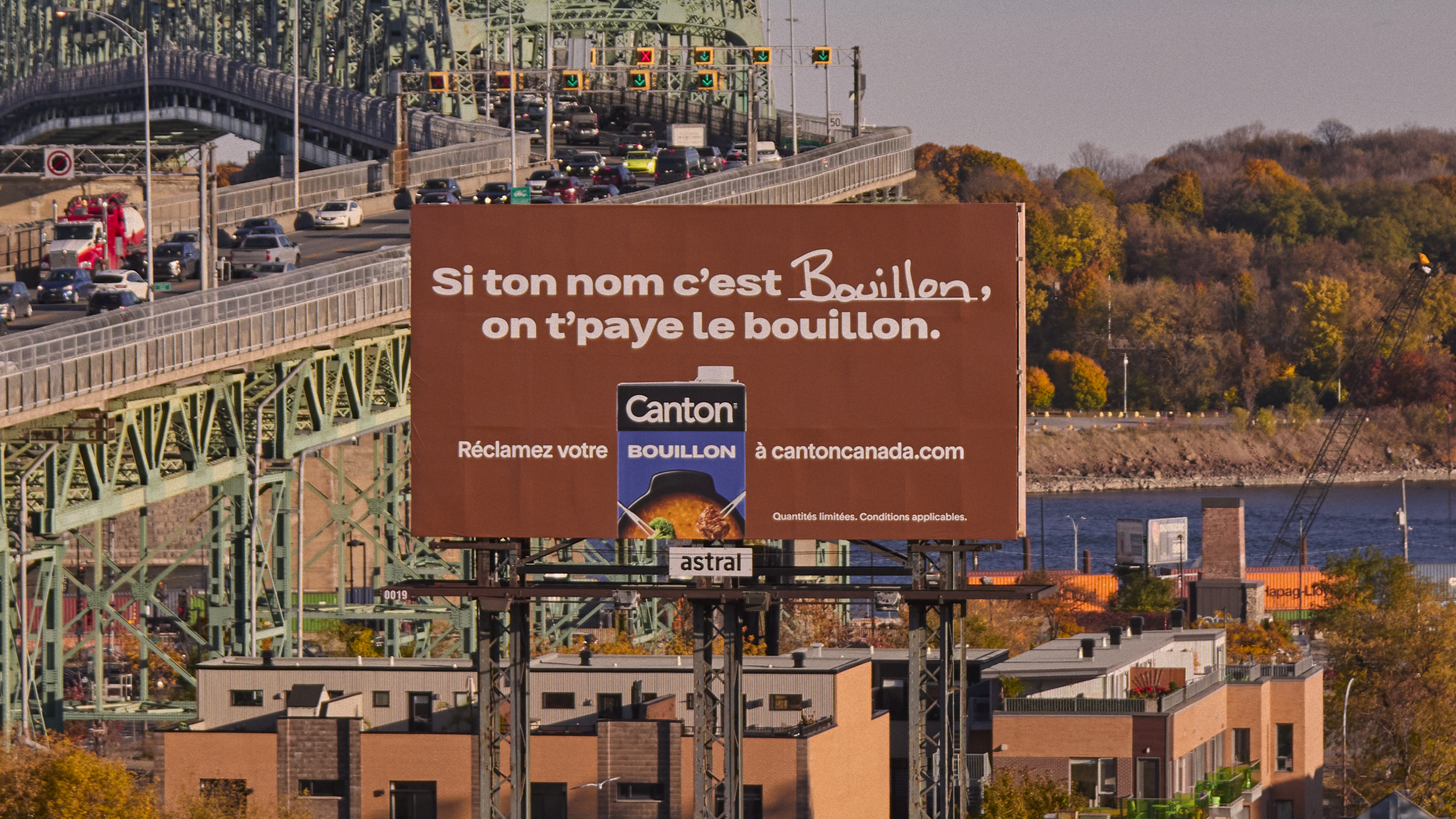 Une grande publicité sur une panneau d'affichage en français. Le panneau dit : « Si ton nom c’est Bouillon, on t'paye le bouillon. » Il y a une image d’une boîte de bouillon de la marque Canton avec un bol de soupe. En arrière-plan, il y a un pont avec beaucoup de voitures et une rivière, ainsi que des arbres aux couleurs automnales.