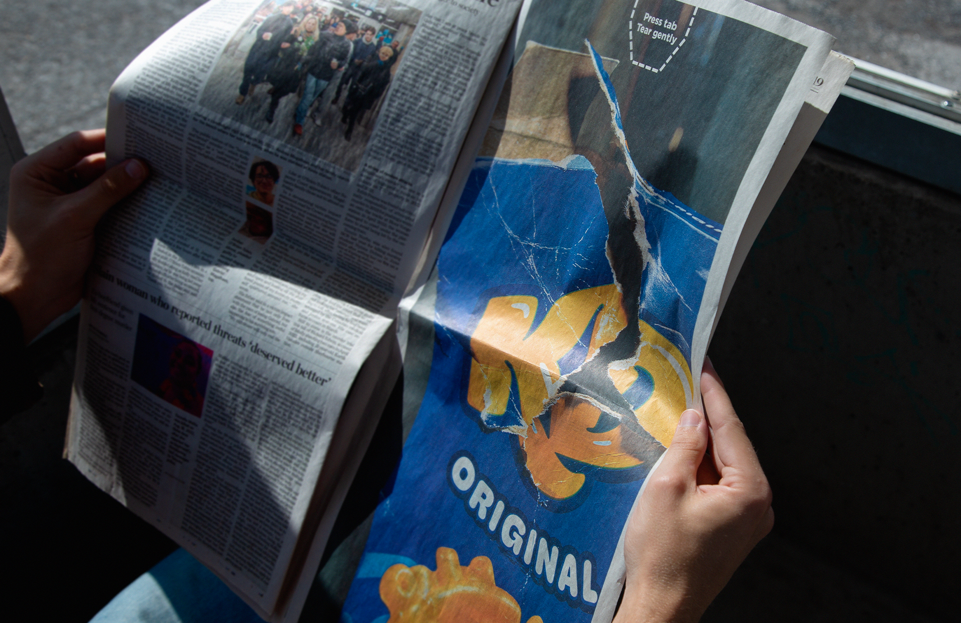 Une personne tient un journal ouvert dans une main et un emballage de chips dans l'autre. Le journal montre une photo de groupe et un article, tandis que l'emballage de chips est bleu avec une illustration d'une tête de dinosaure jaune et l'inscription "ORIGINAL".