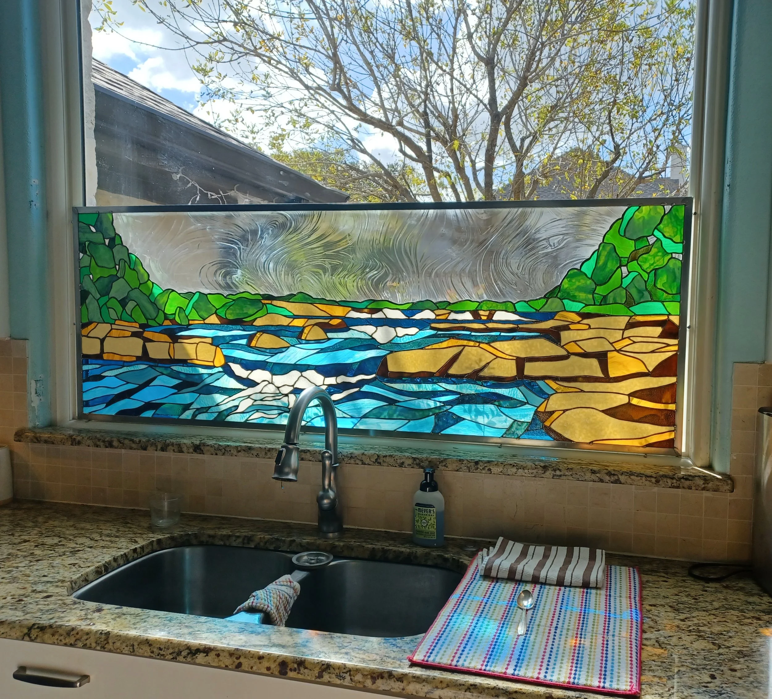 Custom stained glass installed behind residential kitchen sink, partially blocking the view while allowing light and color to enter