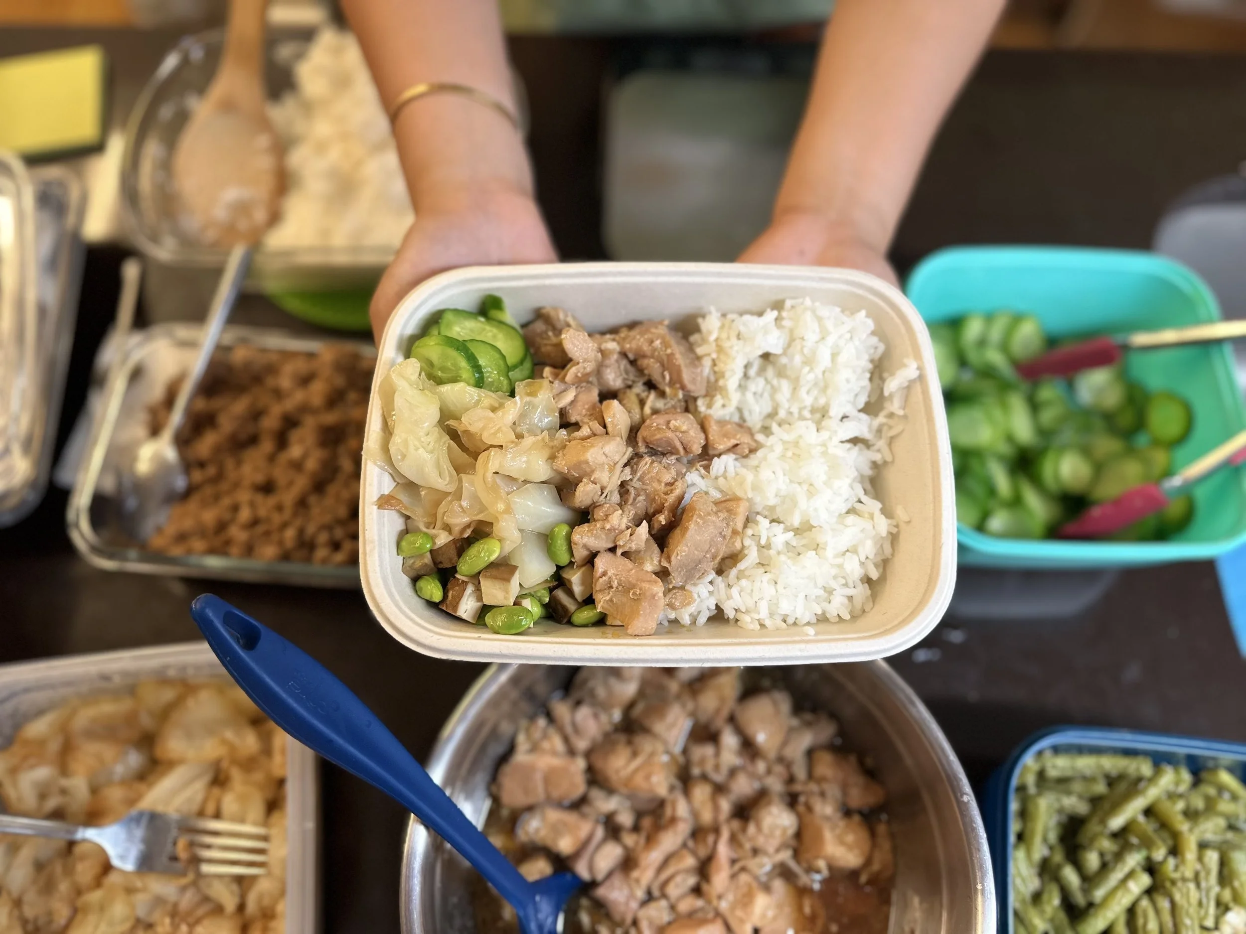 hand holding a meal container with white rice, chopped chicken, and sliced cucumbers, with assorted side dishes surrounding it.