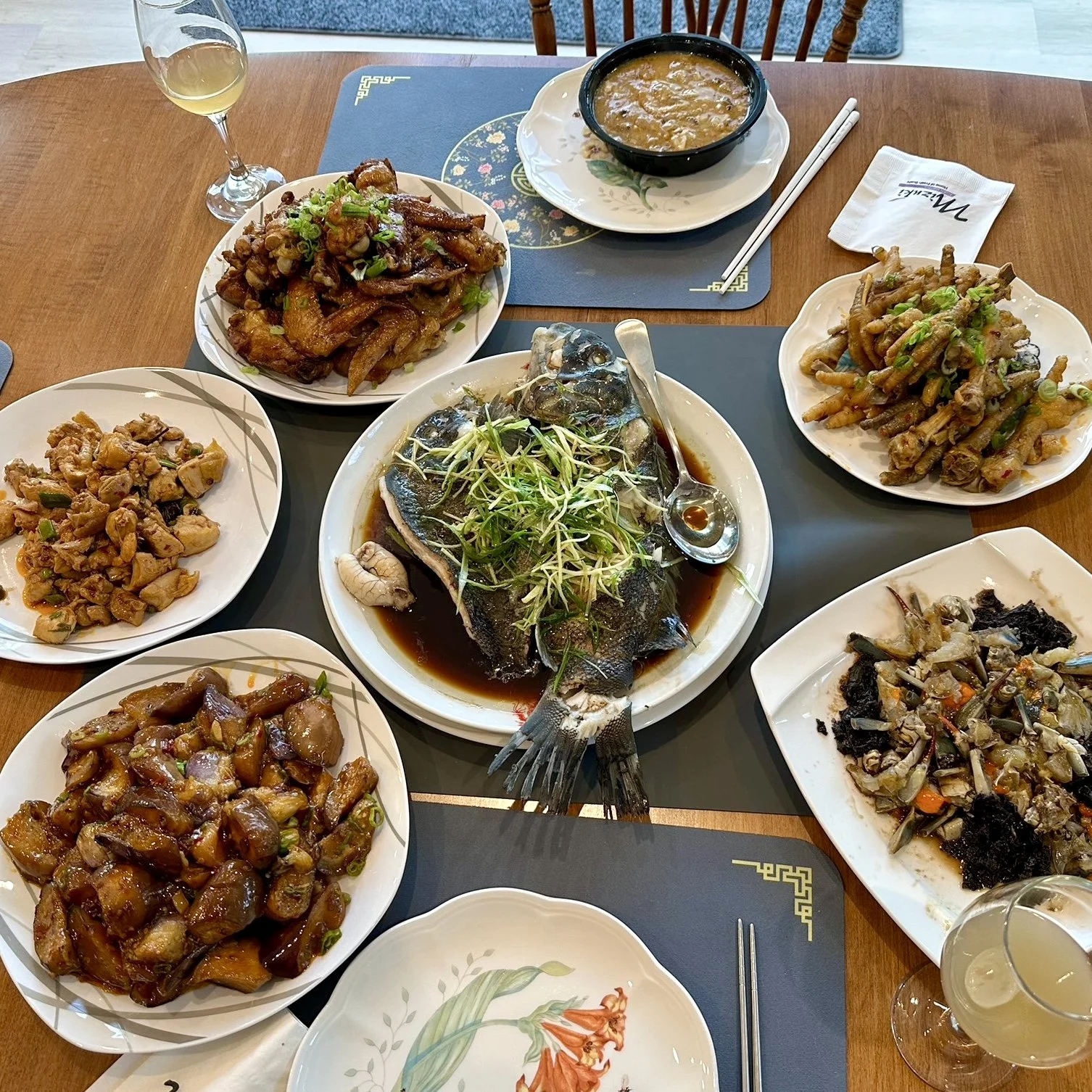 A Chinese seafood dinner with a whole fish in soy sauce garnished with green sprouts, side dishes including stir-fried vegetables, chicken, and an egg drop soup, with glasses of white wine on a wooden table.