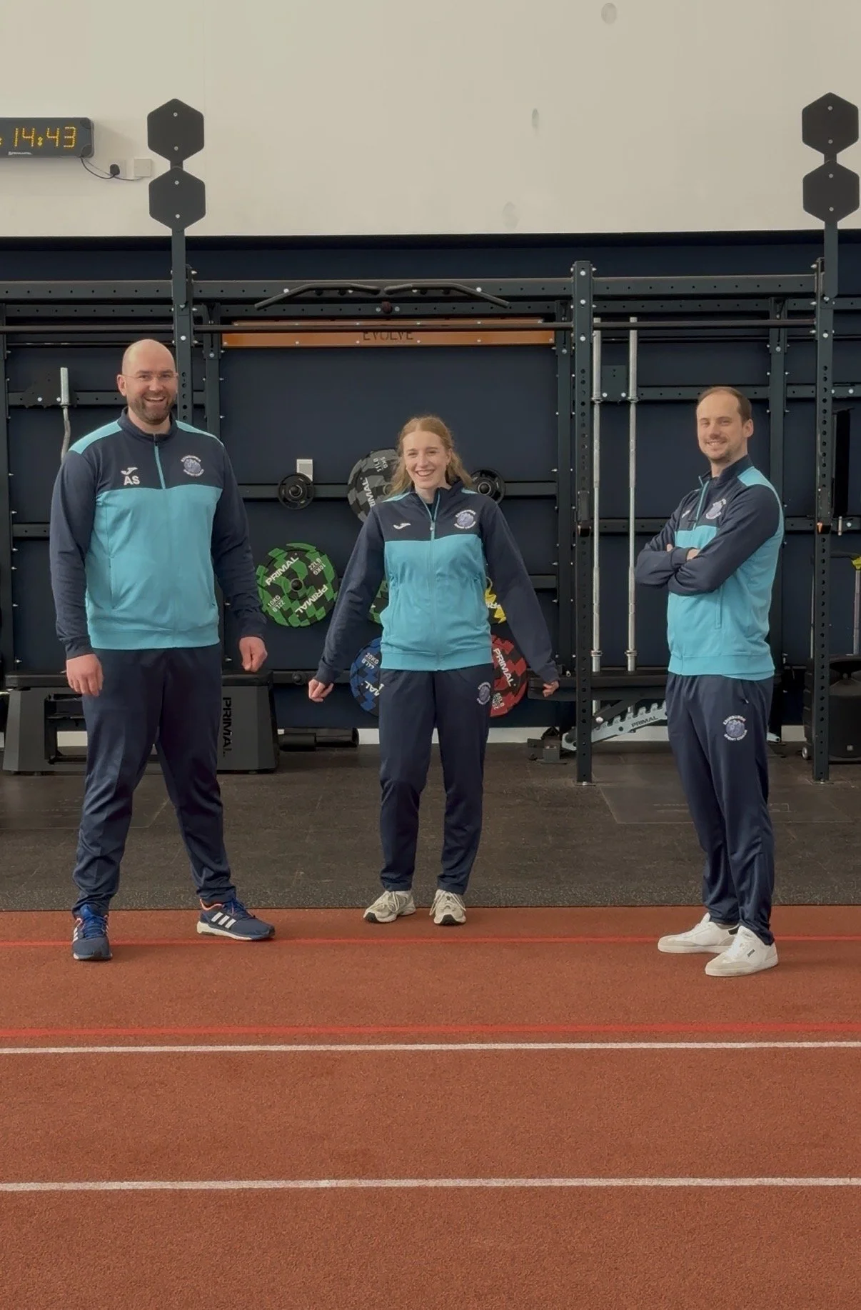 Three athletes in matching blue and navy track suits standing inside a gym. They are smiling and posing for the photo with exercise equipment and a timer in the background.