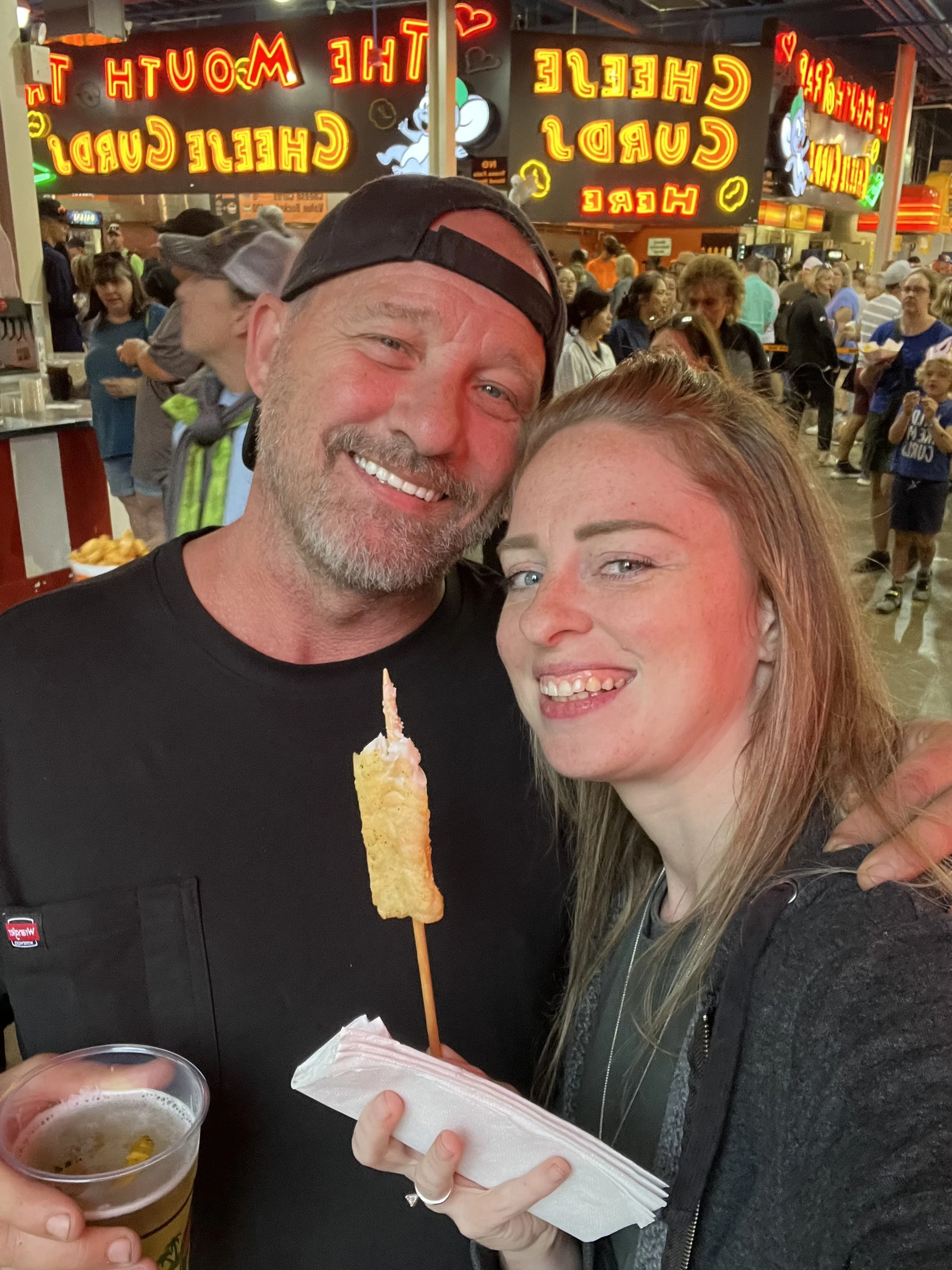 Two smiling people, a man and a woman, at a crowded indoor event, holding a piece of fried food on a skewer and a plastic cup of beer, with bright neon signs in the background.
