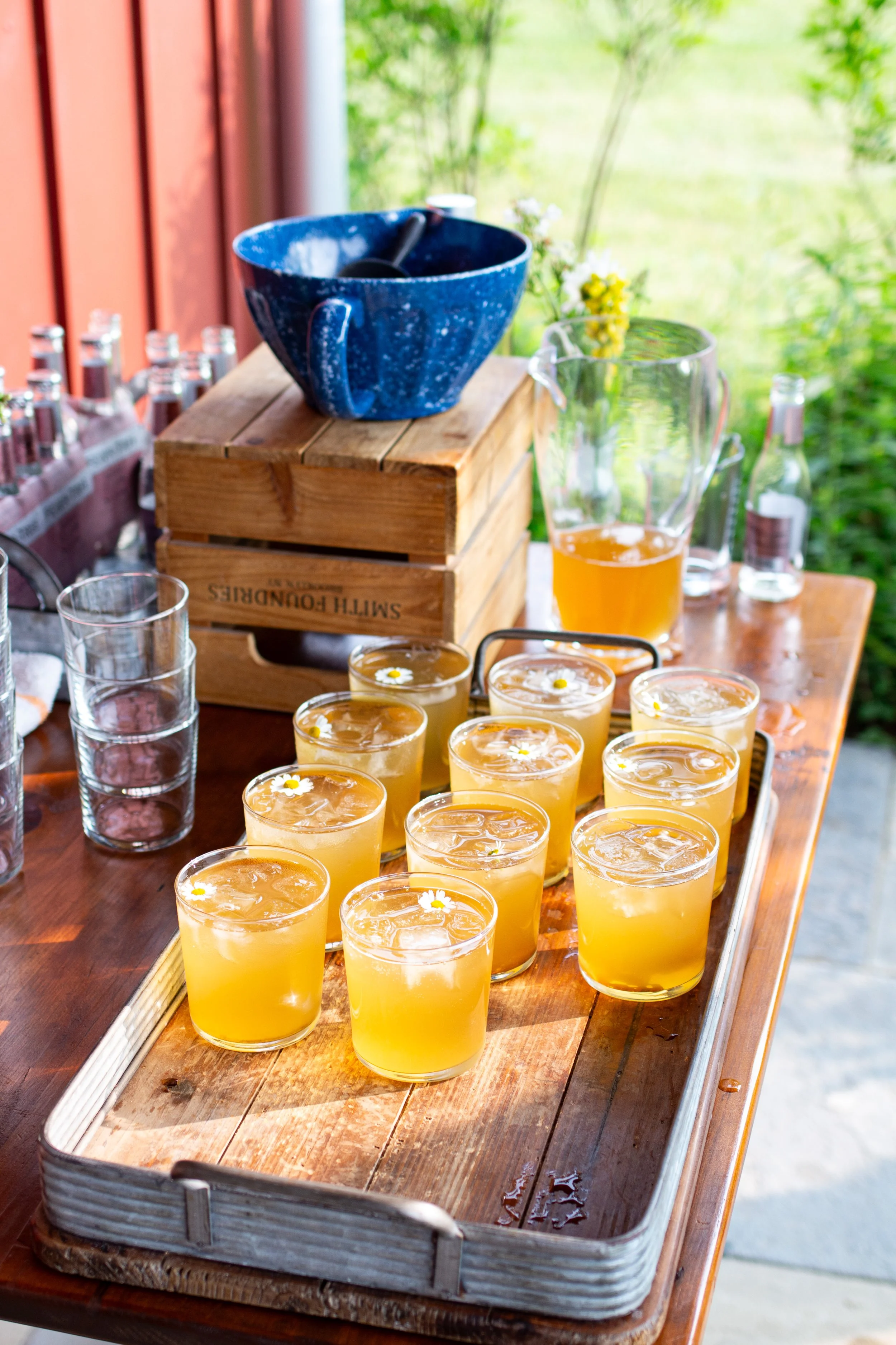 A wooden table holds glasses of yellow juice with small white flower garnishes, a large pitcher, and a blue bowl on a bright outdoor porch with greenery in the background.